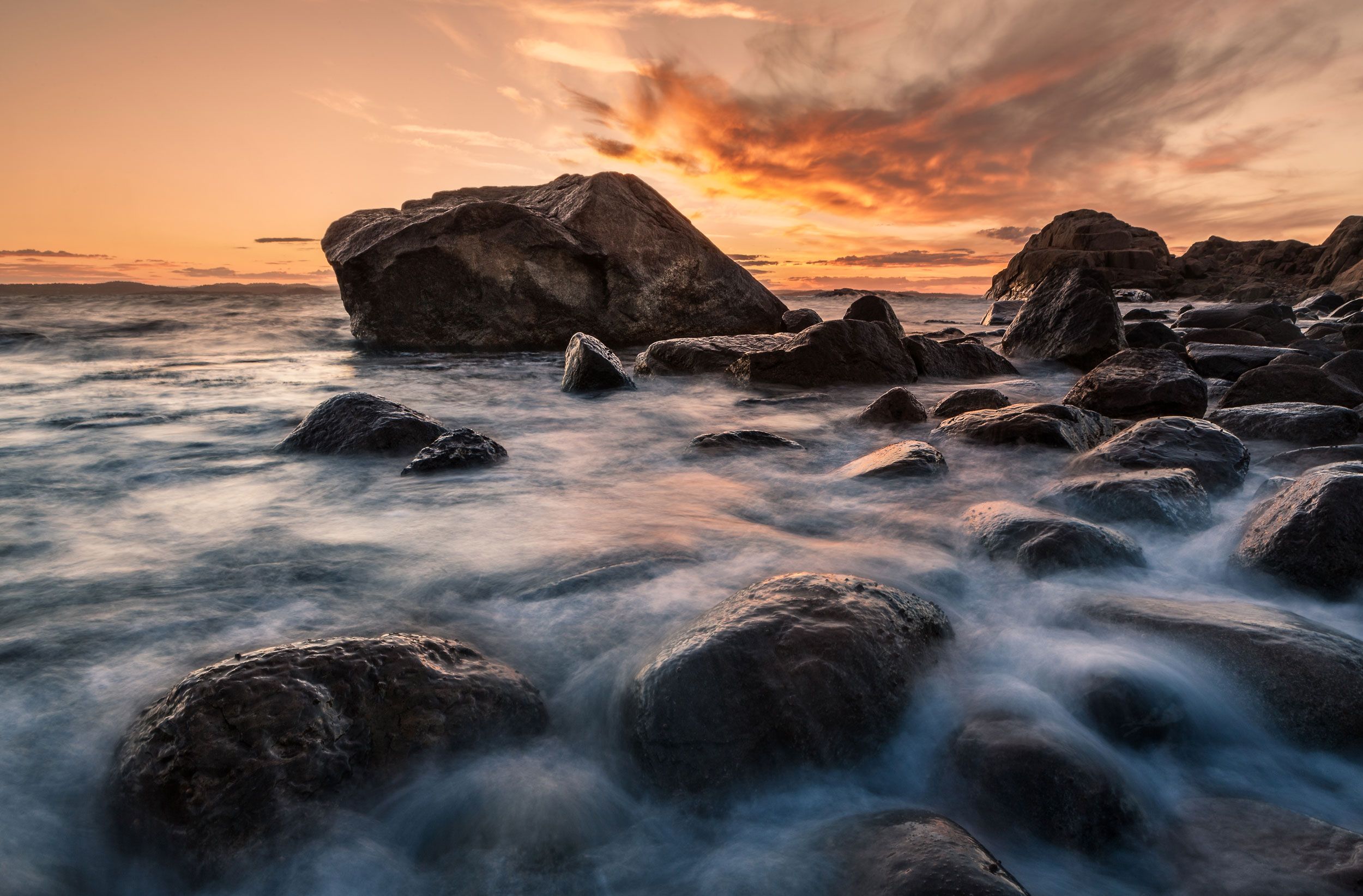 Dramatisk solnedgang over steinete strand med bølger som skyller rundt mørke fjellforminger.