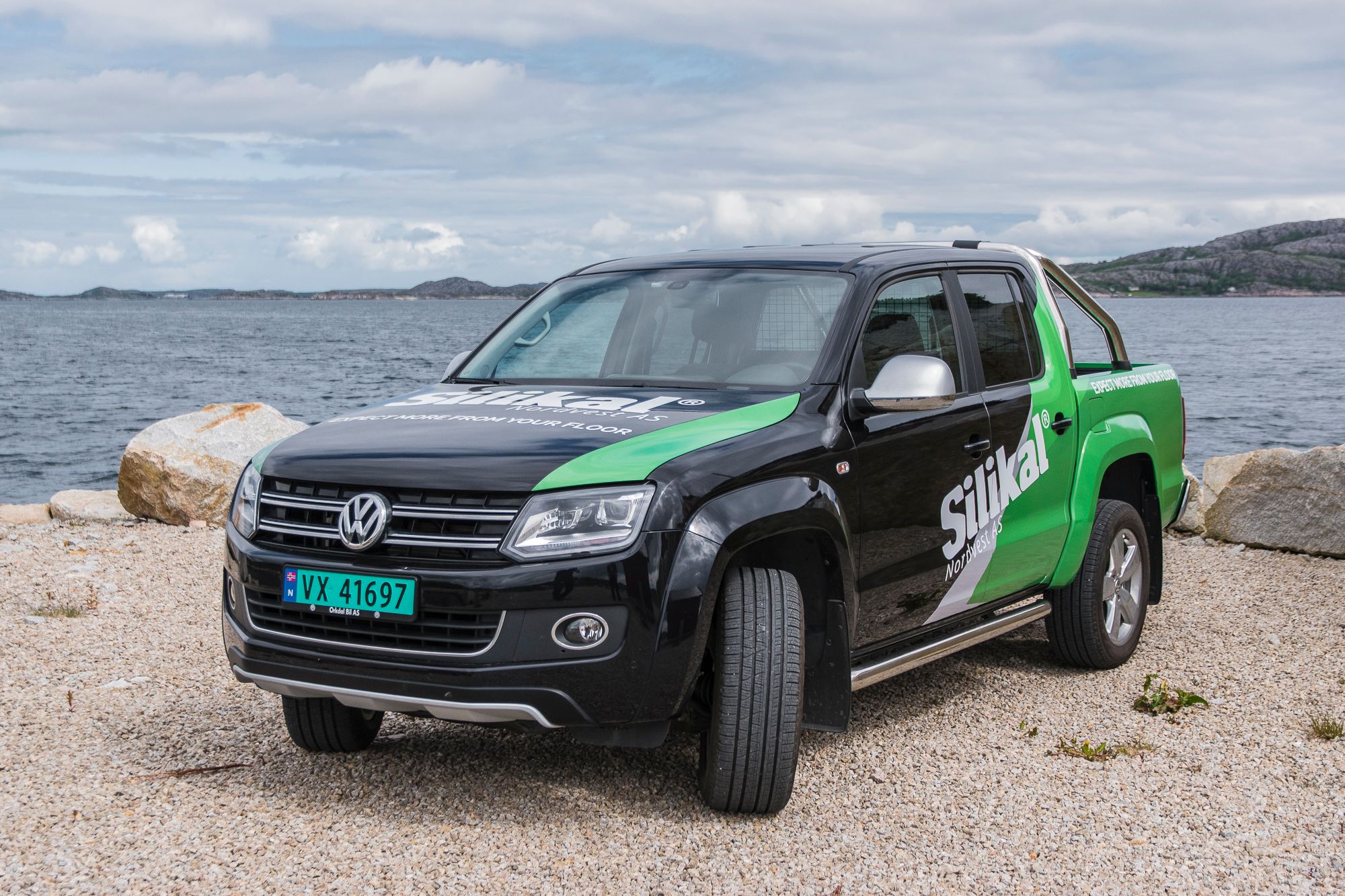 Svart og grønn Volkswagen Amarok pickup med Silikal-logoen parkert på strandsand ved fjorden.