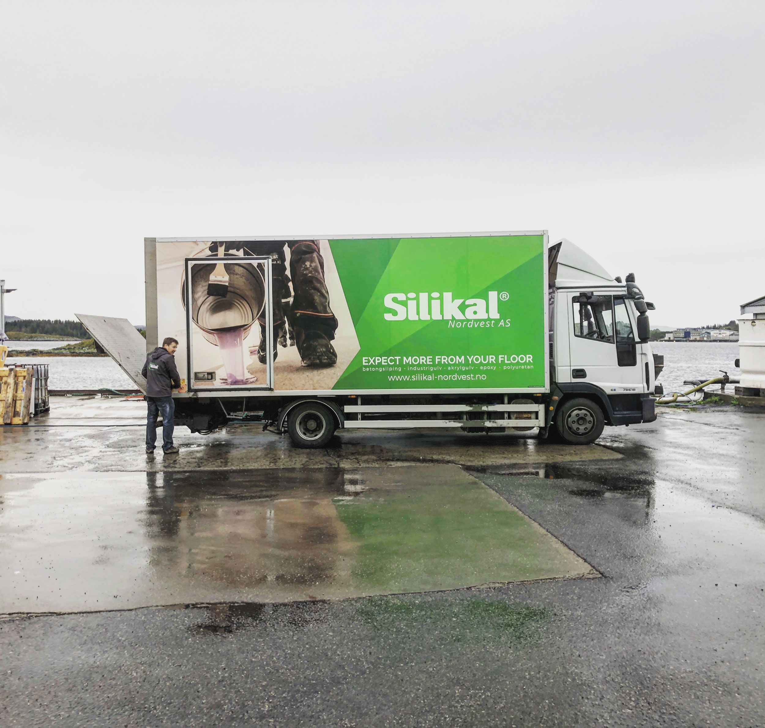 Silikal Nordvest AS lastebil med reklame for industribelegg på våt asfalt, mann står ved siden av kjøretøyet