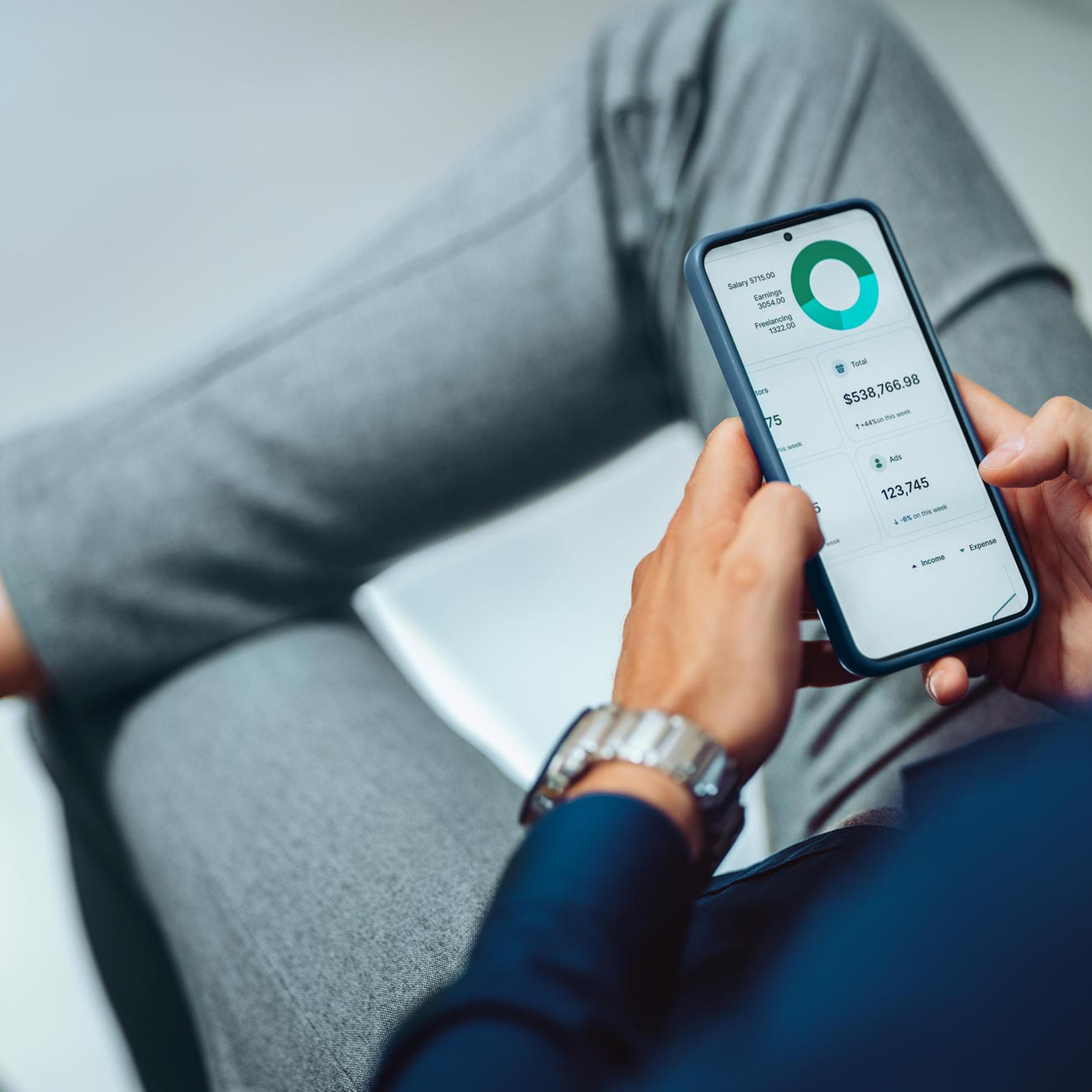Person holder smarttelefon med finansiell dashboardapp som viser sparesamtale, utgifter og økonomisk oversikt.