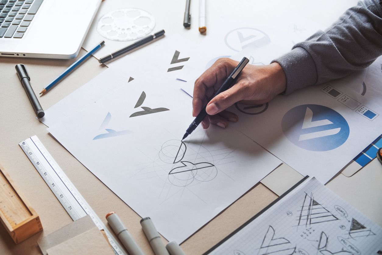 Designer skisserer logodesign på papir med pensler og markører, omgitt av designverktøy og skisser på arbeidsbrettet.