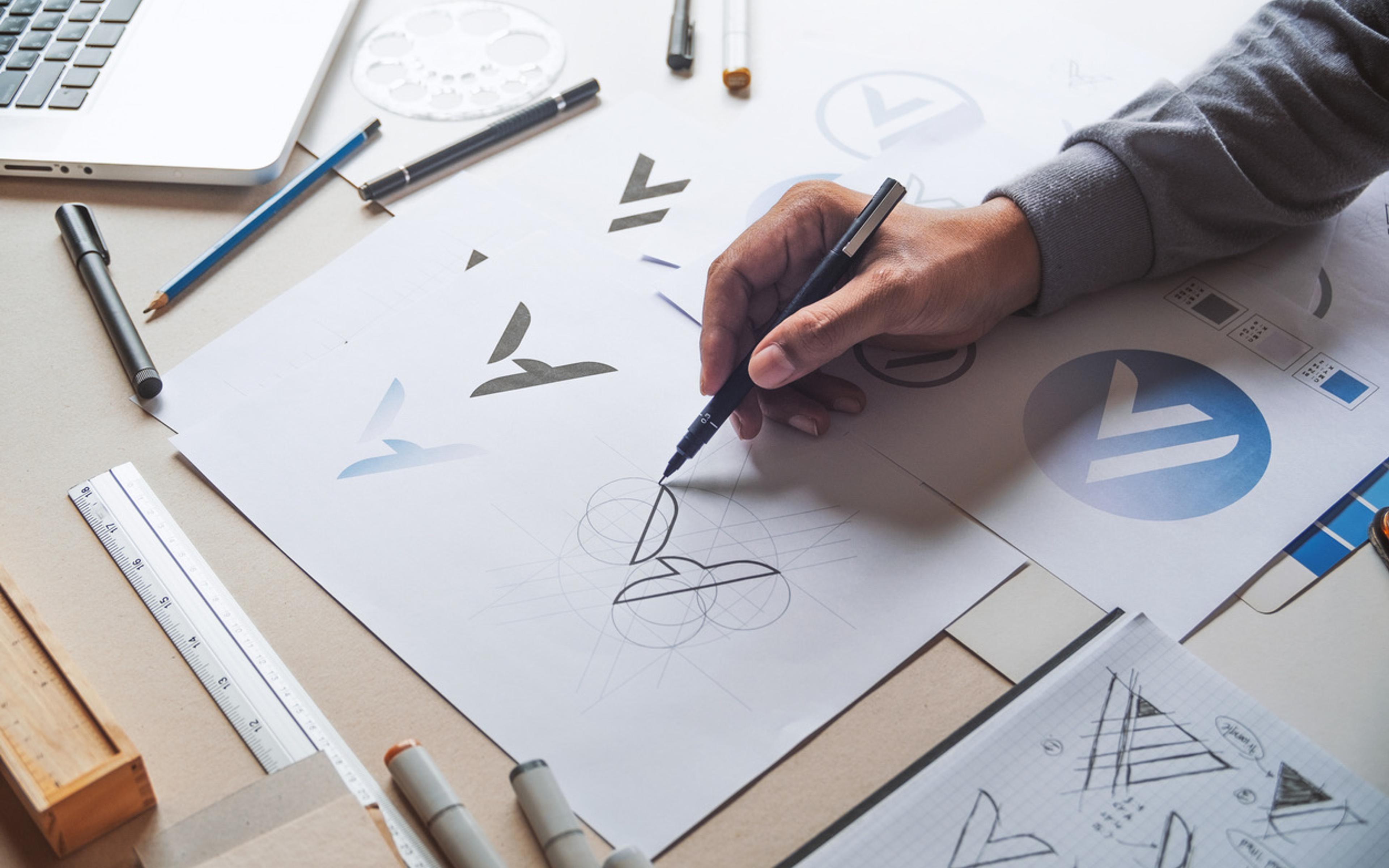 Designer skisserer logodesign på papir med pensler og markører, omgitt av designverktøy og skisser på arbeidsbrettet.