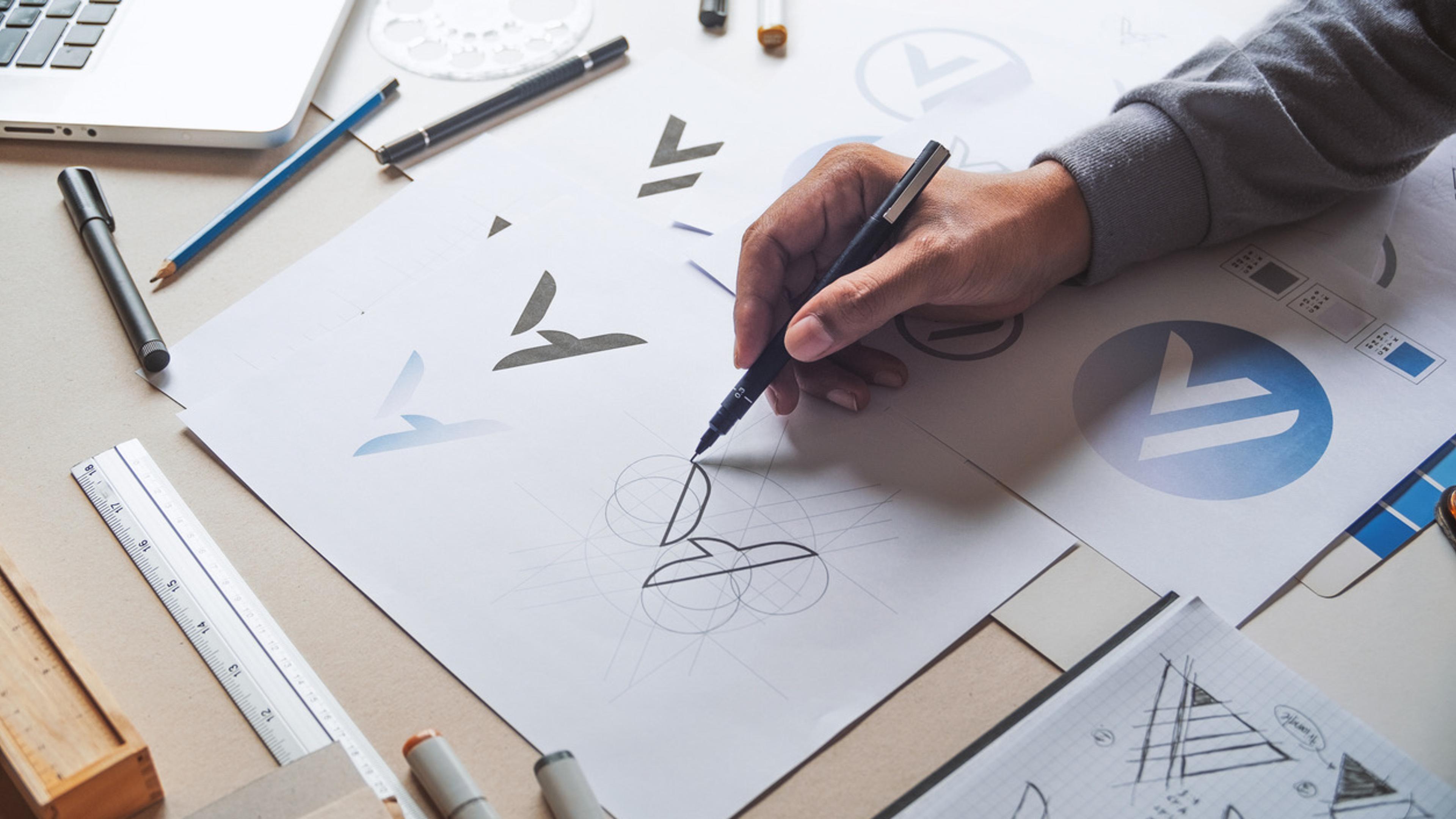 Designer skisserer logodesign på papir med pensler og markører, omgitt av designverktøy og skisser på arbeidsbrettet.