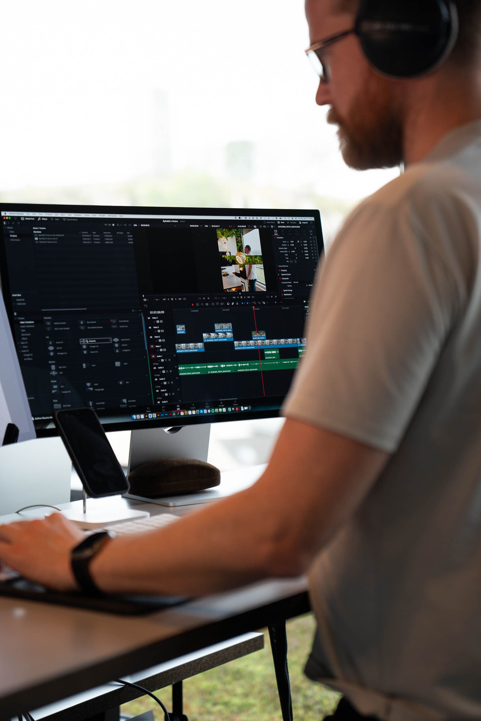 Videoredaktør med hodetelefoner arbeider ved skjerm med videoredigeringsprogram og tidslinjer, med byutsikt i bakgrunnen.