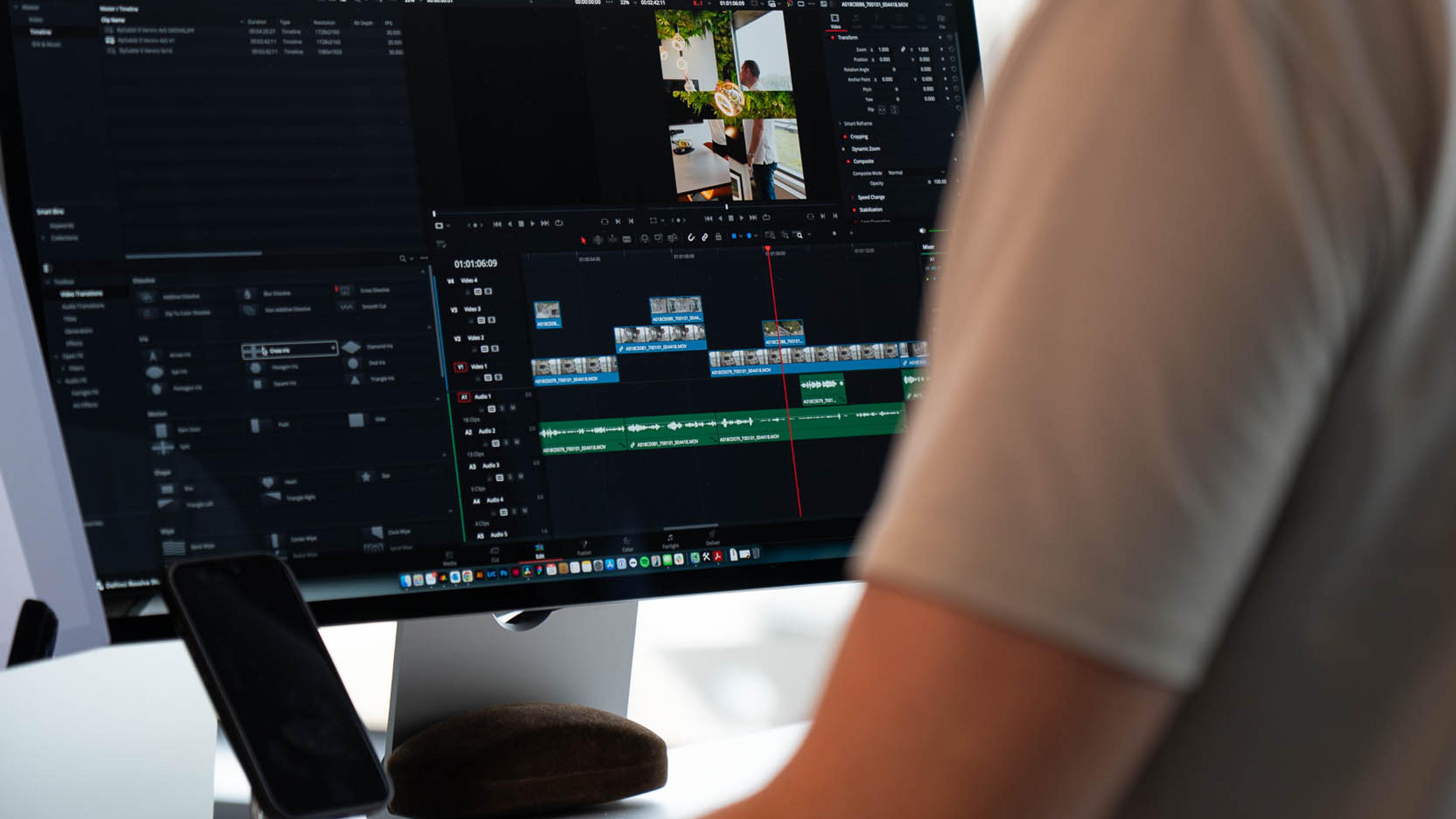 Videoredaktør med hodetelefoner arbeider ved skjerm med videoredigeringsprogram og tidslinjer, med byutsikt i bakgrunnen.