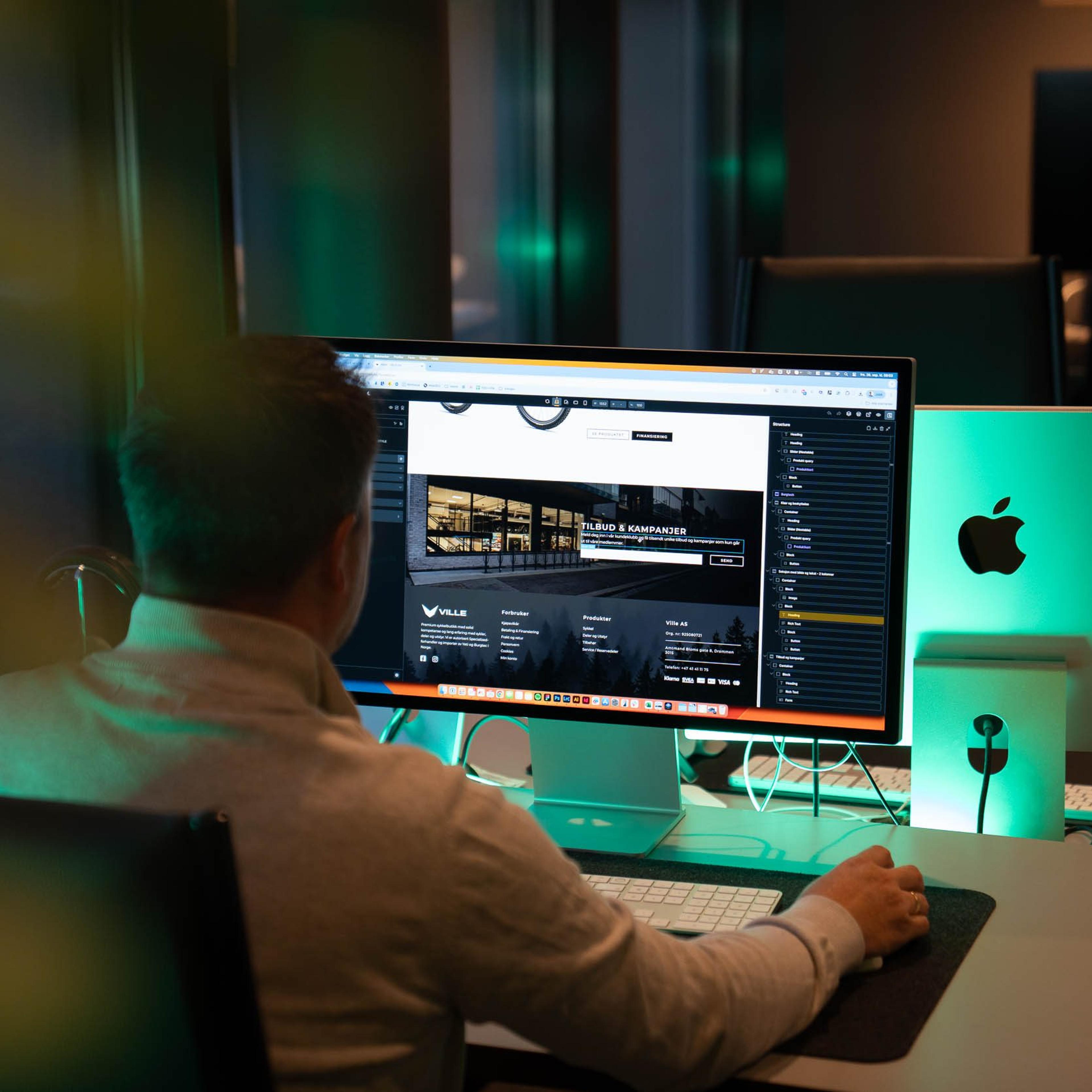 Webutvikler jobber med en nettside på en stor skjerm ved siden av en iMac på kontoret.