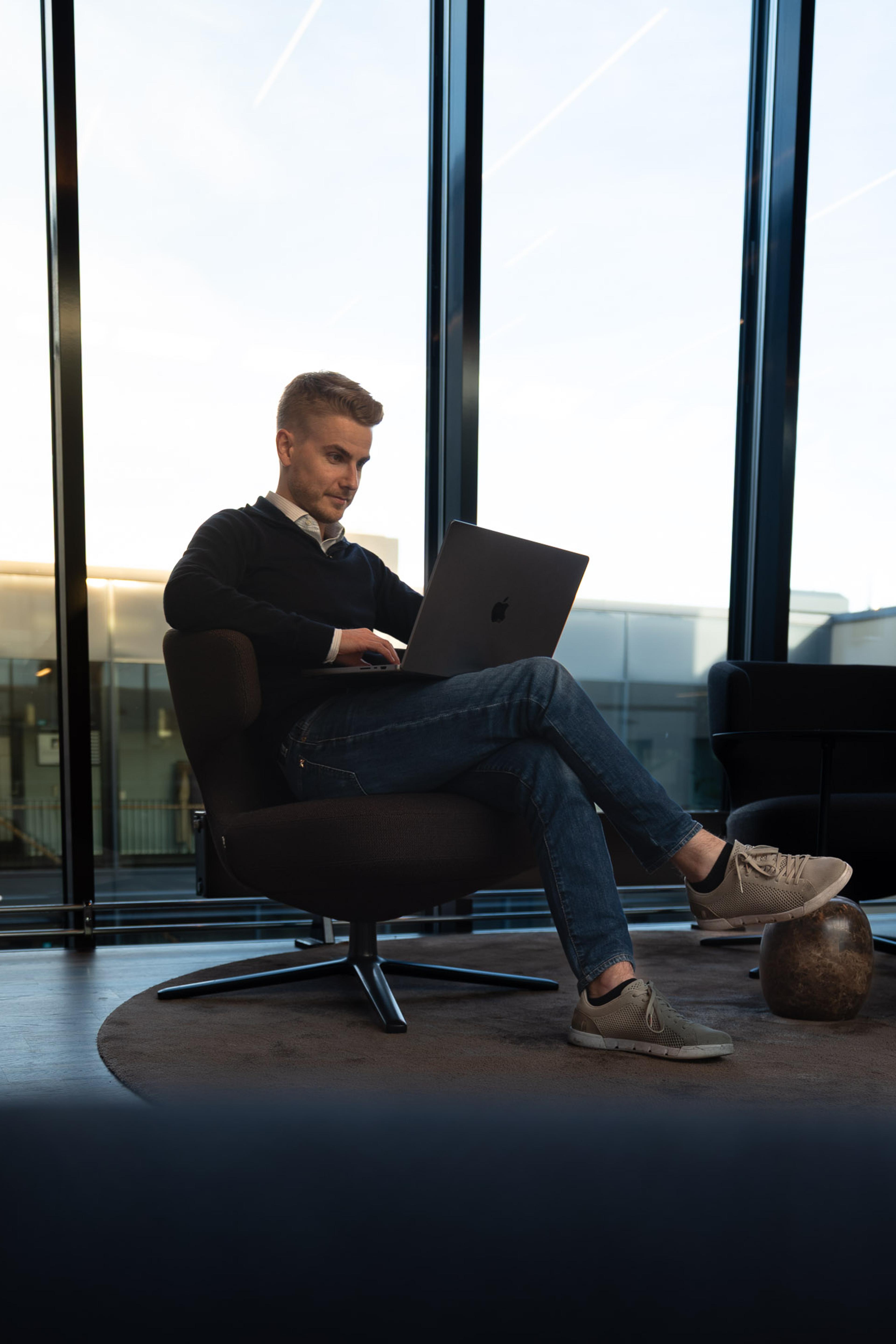 Mann i svart genser arbeider på laptop i moderne kontorlandskap med store vinduer og utsikt.