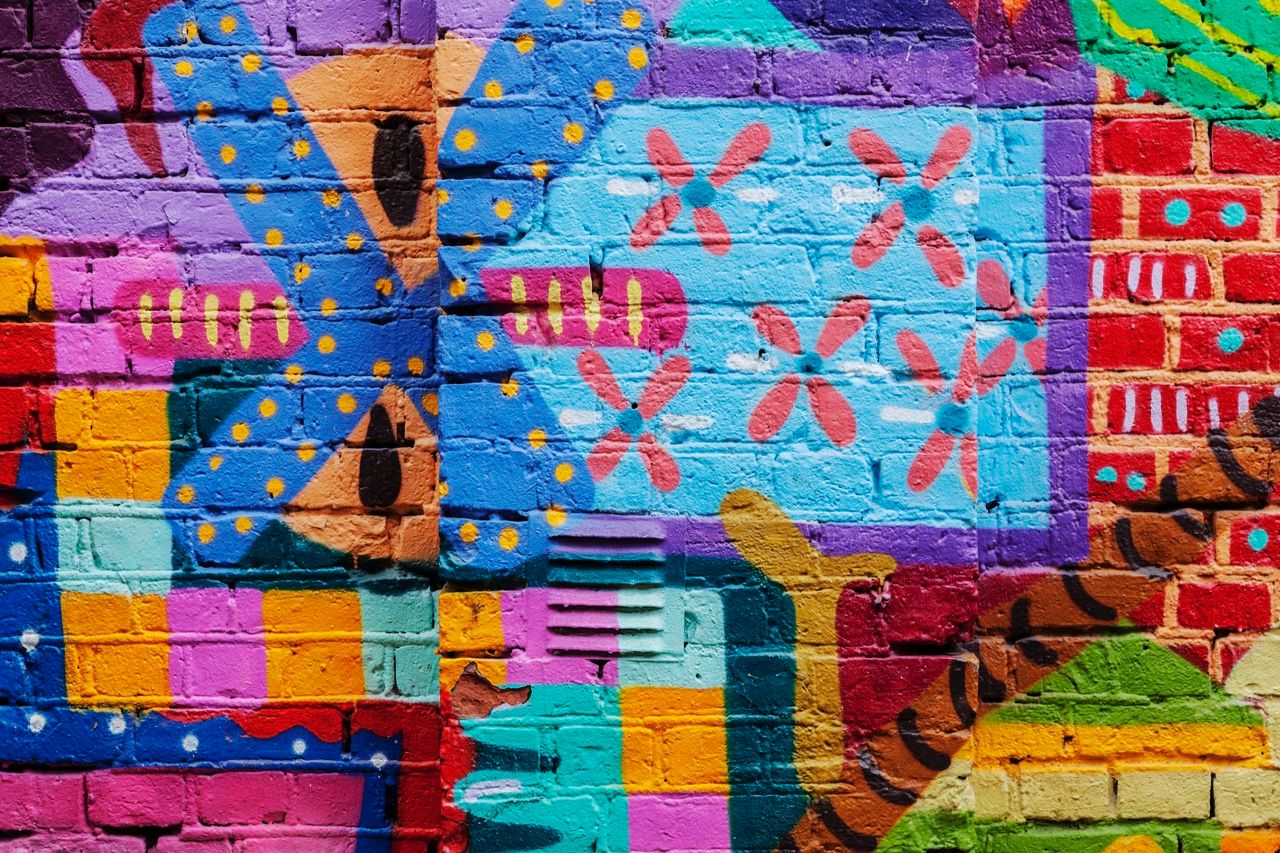 Fargerik street art på murvegg med geometriske mønster, blomster og abstrakte former i blå, rosa, gul, grønn og rød.