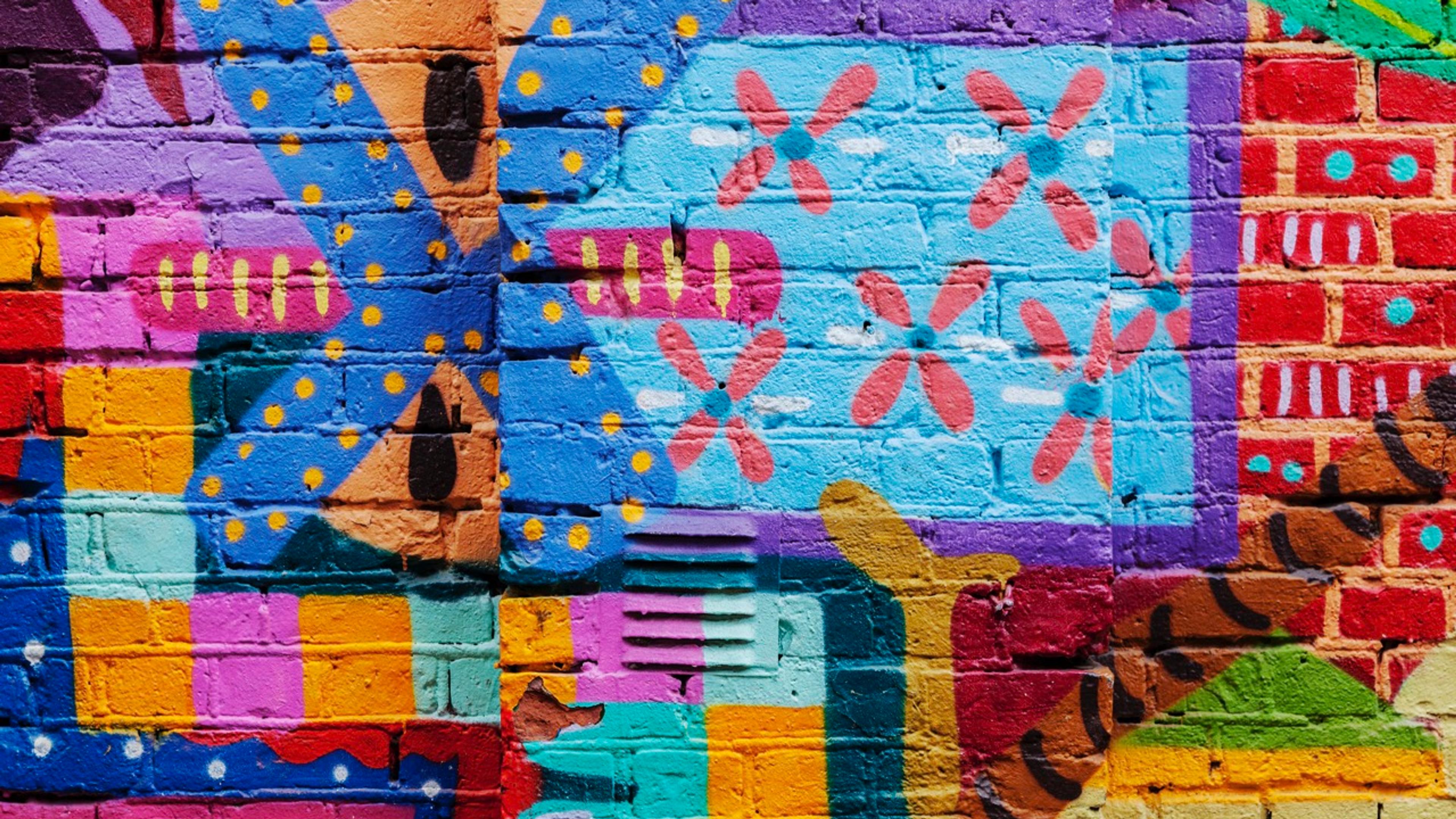 Fargerik street art på murvegg med geometriske mønster, blomster og abstrakte former i blå, rosa, gul, grønn og rød.