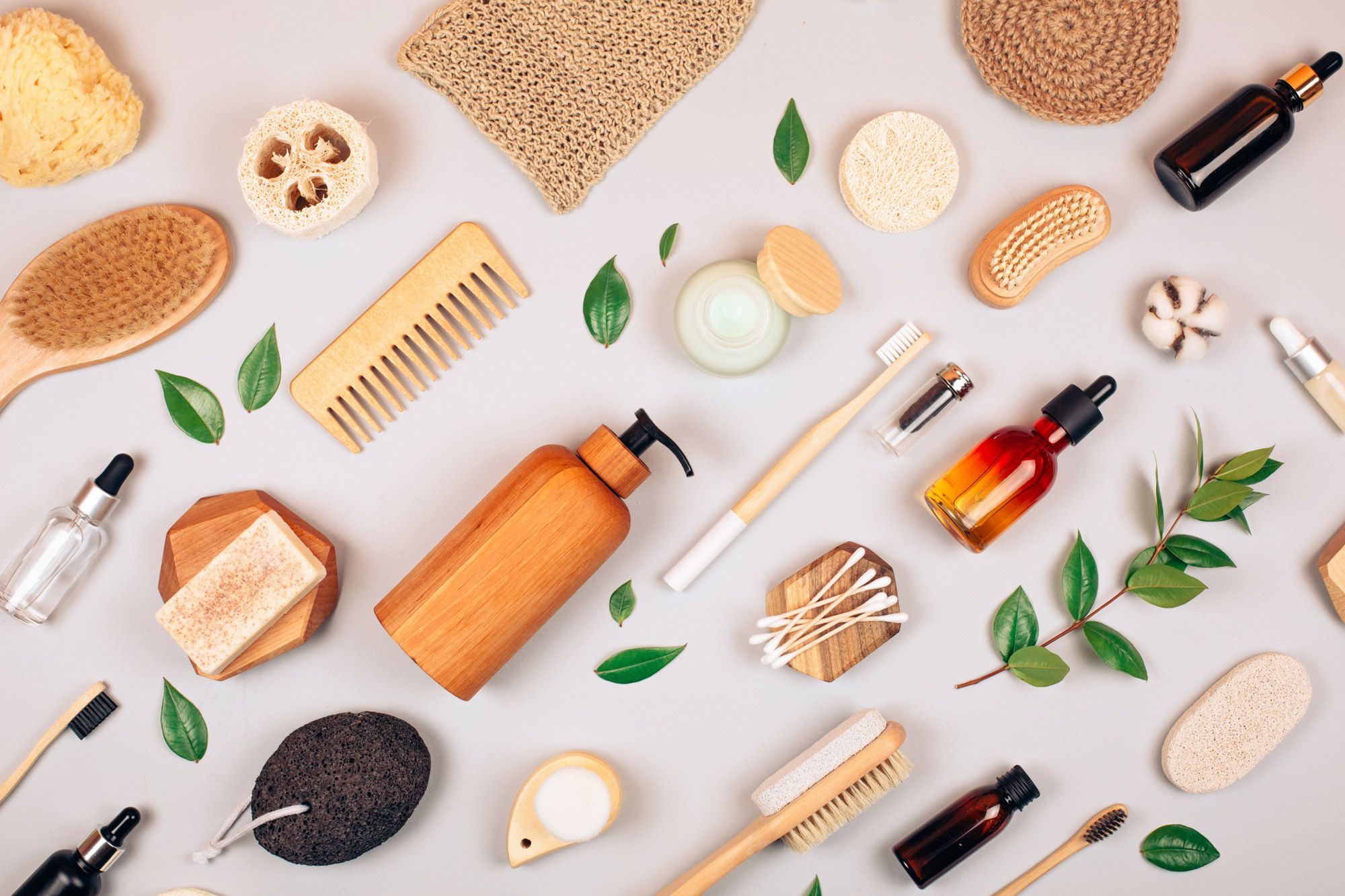 Flat lay av naturlige skjøtselsprodukter med børster, svamper, kremer og essensielle oljer på grå bakgrunn.