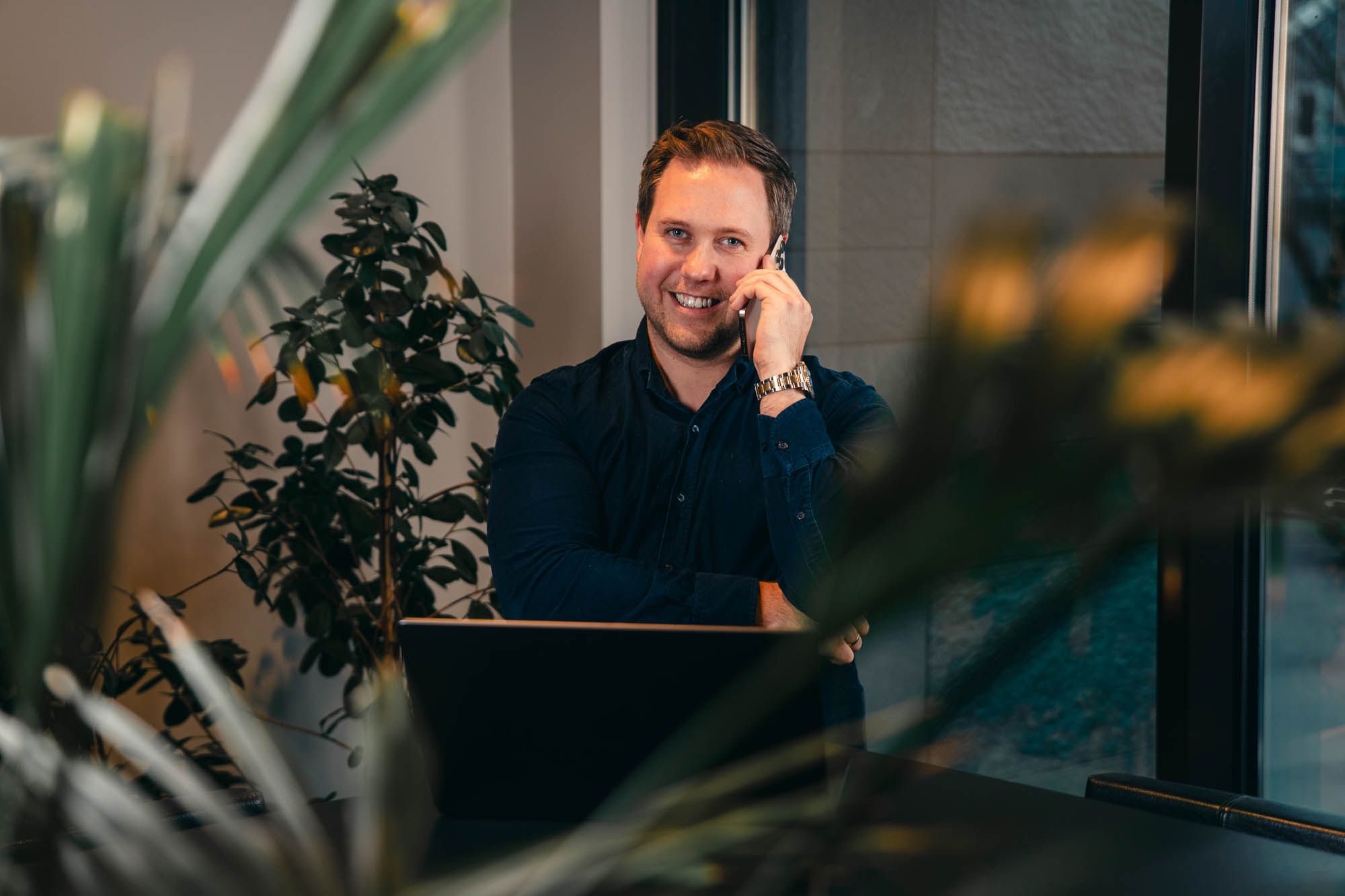 Mann i mørk skjorte smiler mens han tar telefonsamtale ved skrivebord med planter og datamaskinen rundt seg.
