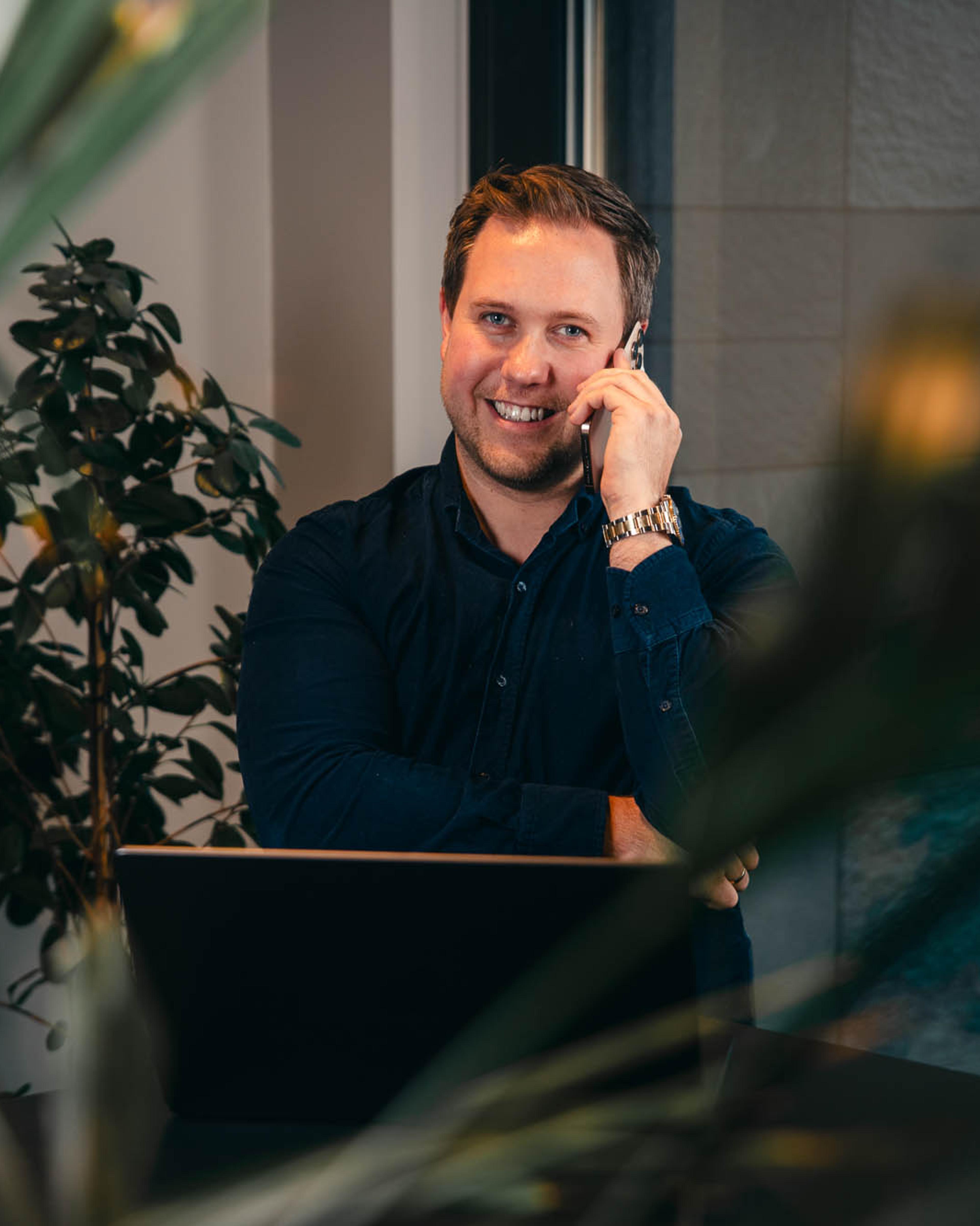 Mann i mørk skjorte smiler mens han tar telefonsamtale ved skrivebord med planter og datamaskinen rundt seg.