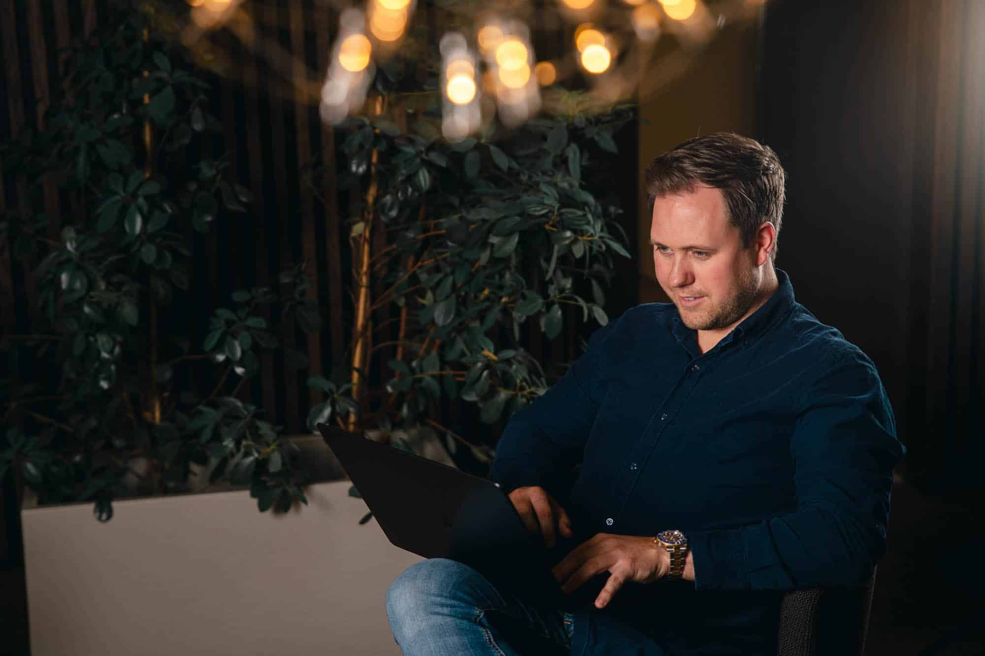 Mann i mørk skjorte arbeider på laptop, omgitt av grønne planter og varm belysning i moderne kontormiljø.