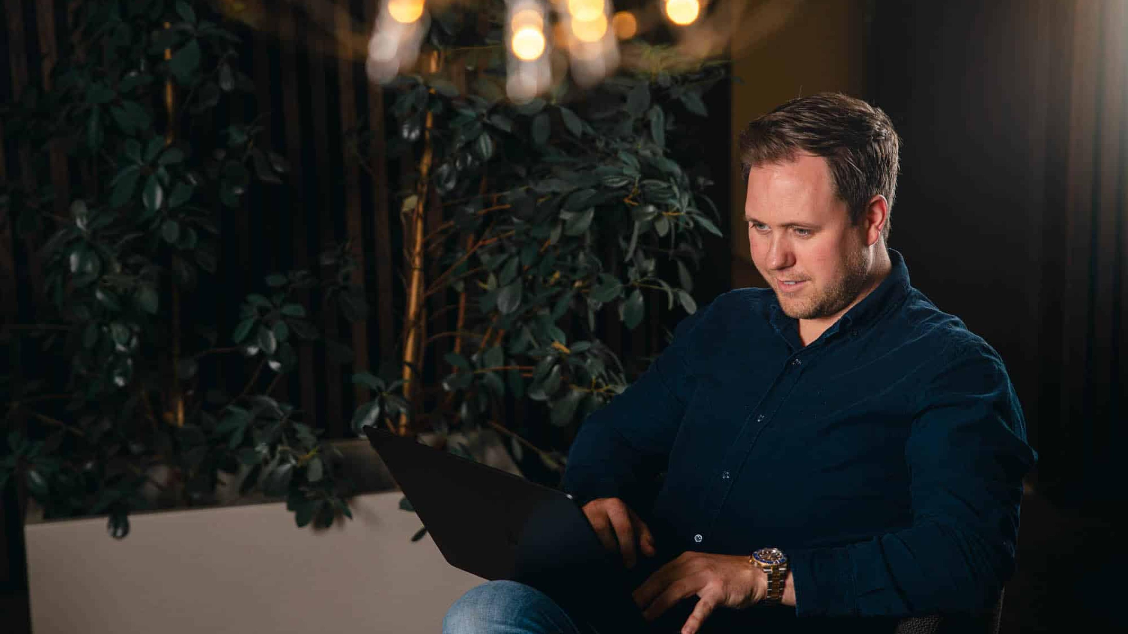 Mann i mørk skjorte arbeider på laptop, omgitt av grønne planter og varm belysning i moderne kontormiljø.