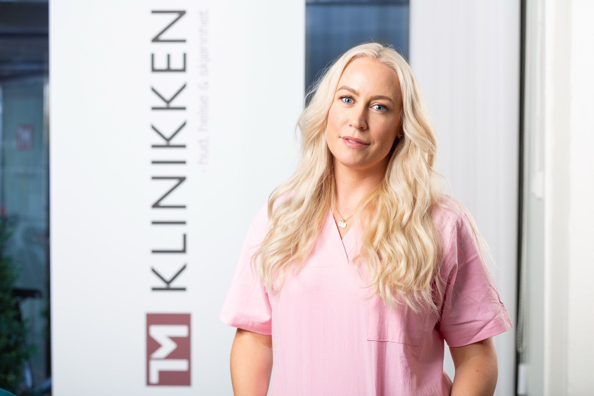 Kvinne med langt blondt hår i rosa uniform står foran KLINIKKEN-logoen på klinikkvegg.