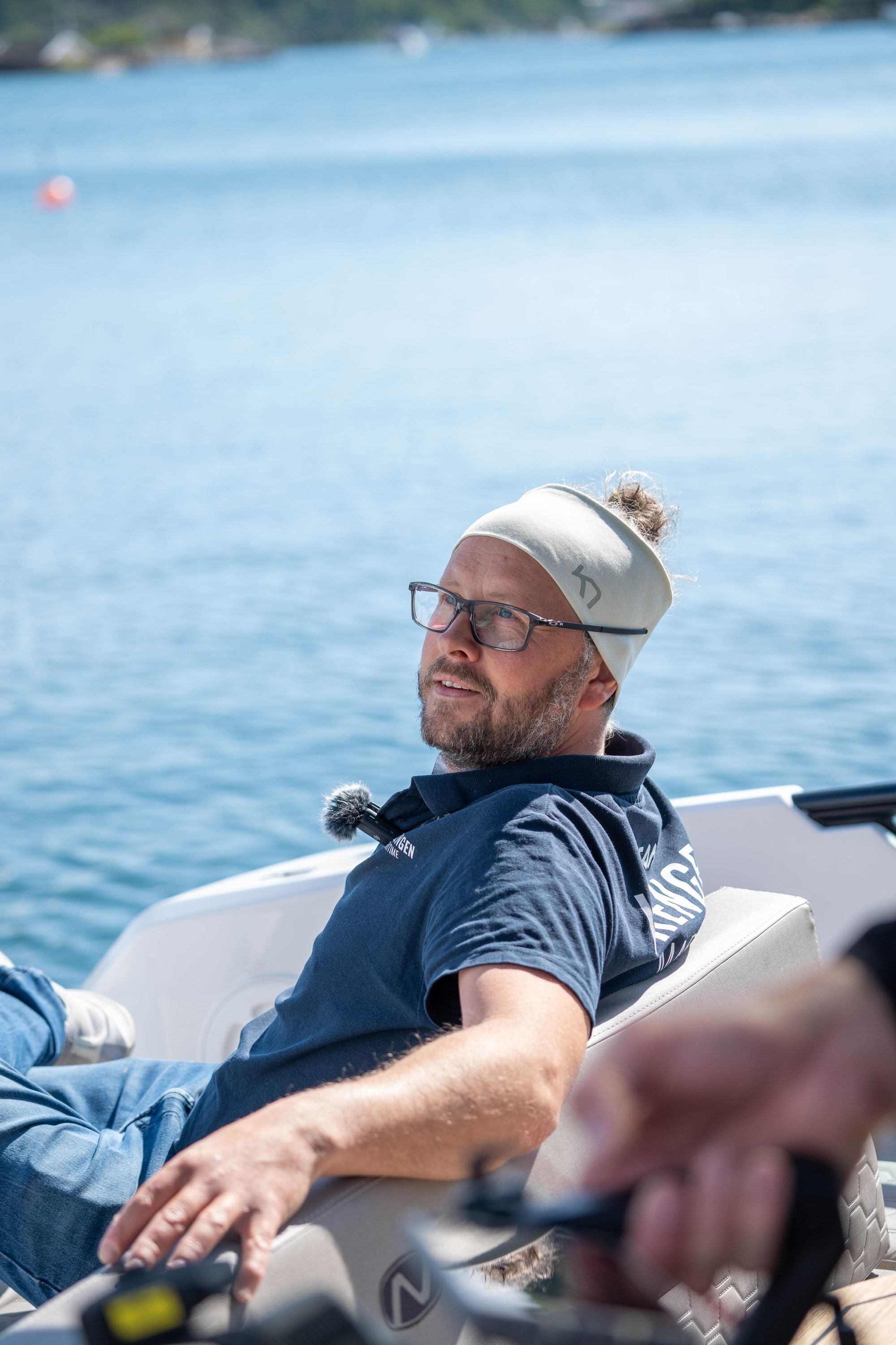 Mann med briller og hvit cap på båt, ser mot kamera med smilende ansikt og casual påkledning.
