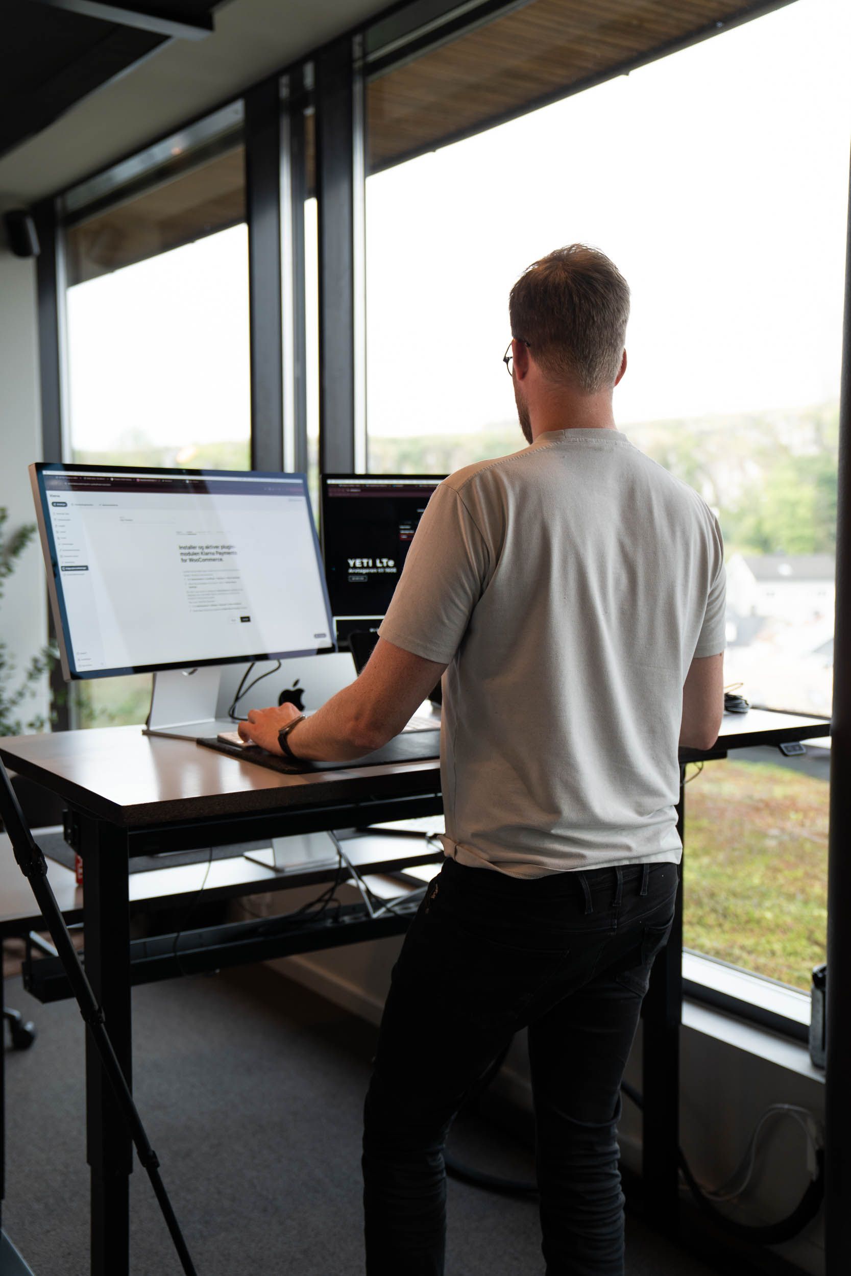 Mann står ved stående skrivebord med monitor og arbeider på WordPress-nettsted med utsikt over landskap gjennom store vinduer.