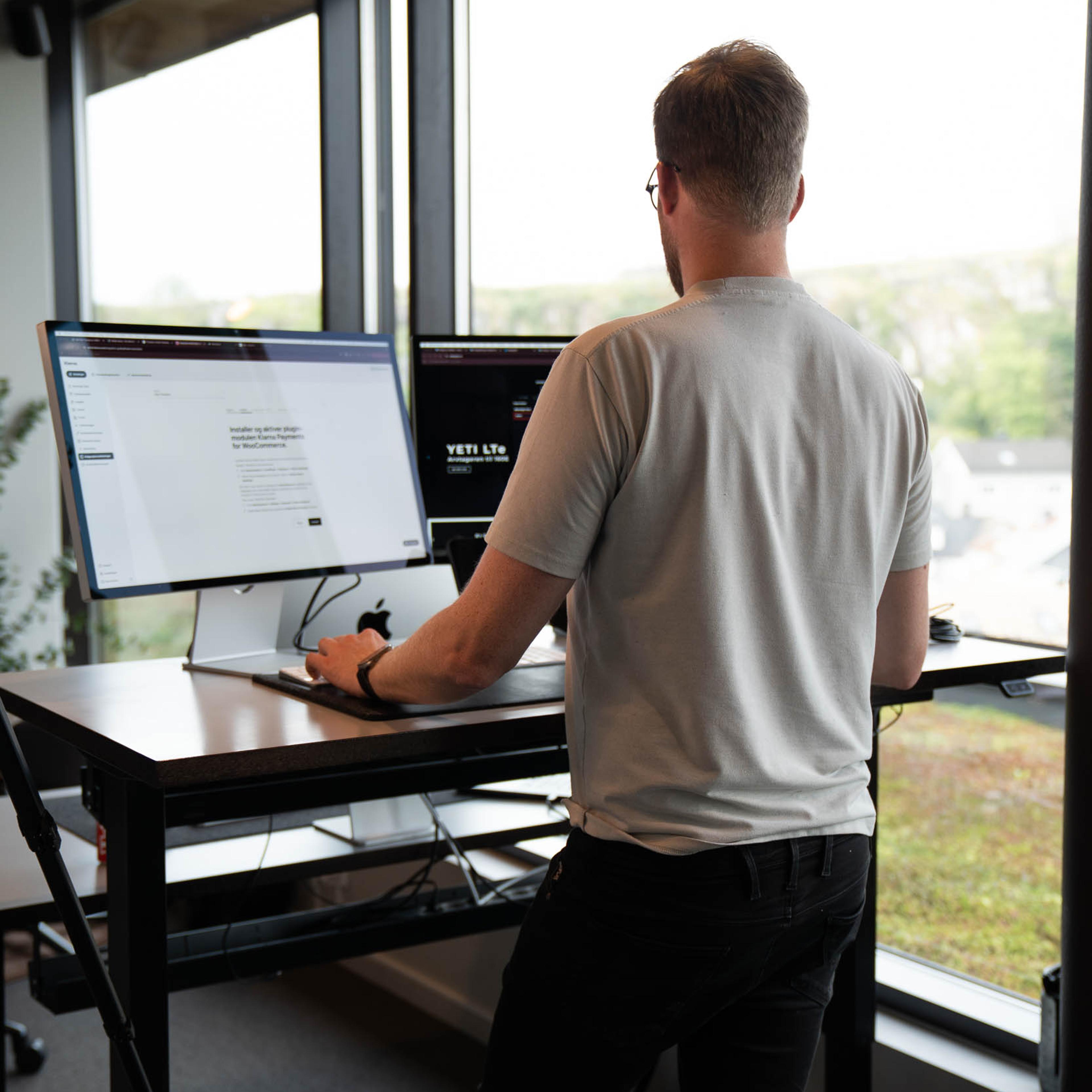 Mann står ved stående skrivebord med monitor og arbeider på WordPress-nettsted med utsikt over landskap gjennom store vinduer.