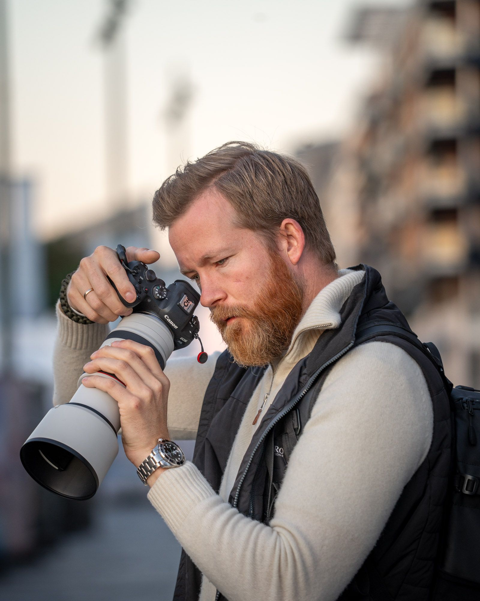 Mann med skjegg fotograferer med profesjonelt kamera og teleobjektiv i urbant miljø med bygninger i bakgrunnen.