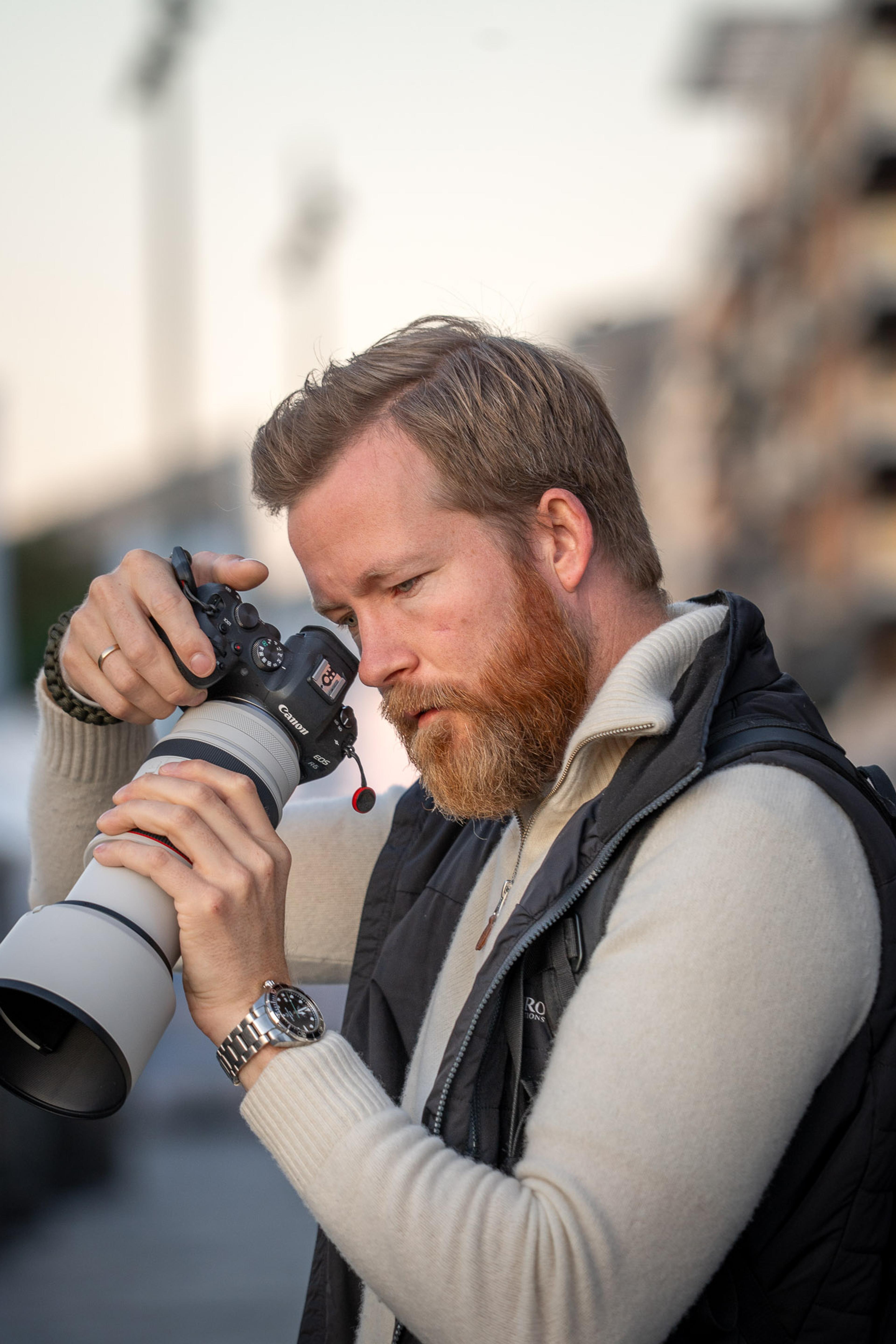 Mann med skjegg fotograferer med profesjonelt kamera og teleobjektiv i urbant miljø med bygninger i bakgrunnen.