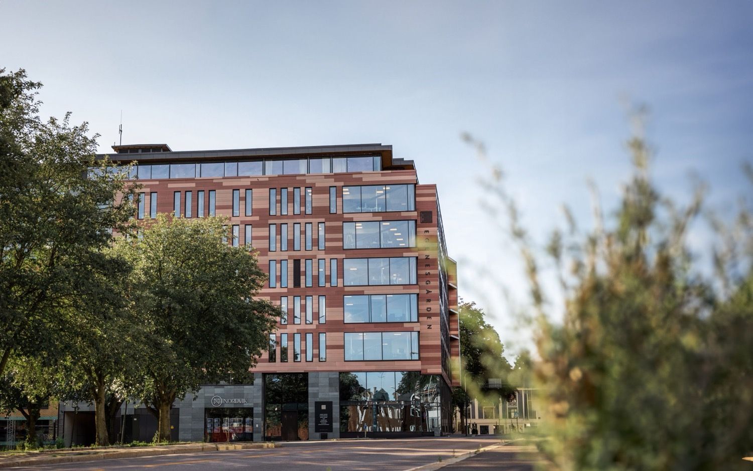 Moderne rødbygning med glassfasade og butikklokaler i Tønsberg sentrum, omgitt av grøntanlegg og trær.