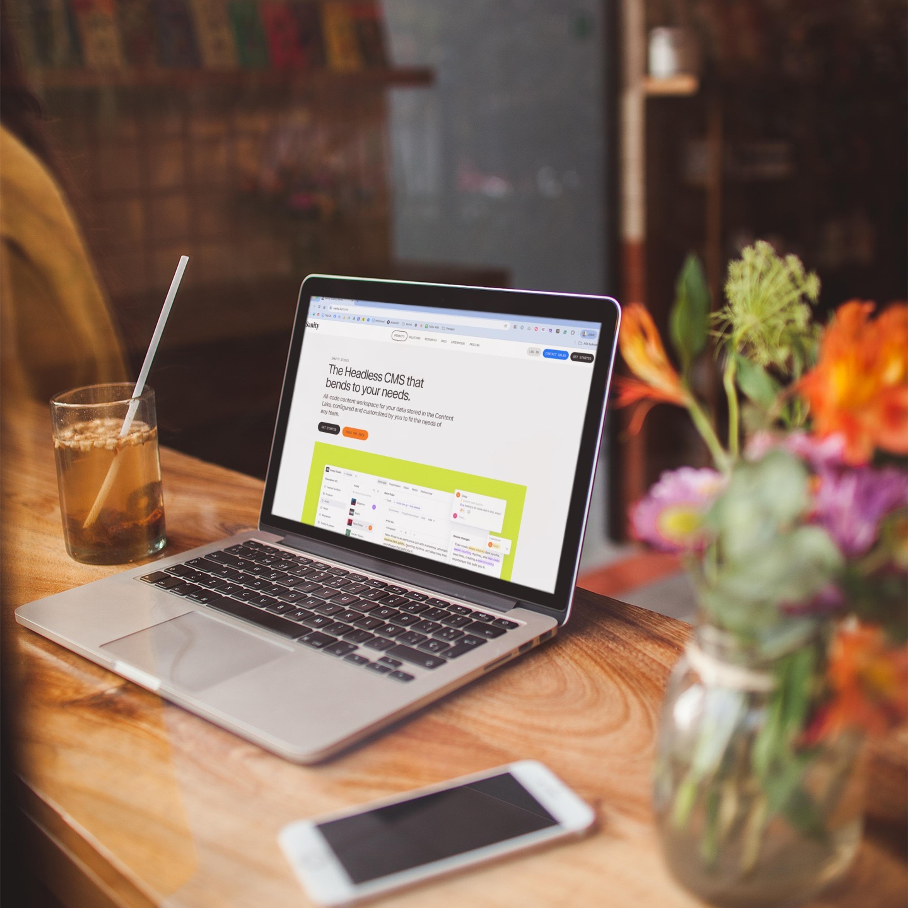 Laptop viser "The Headless CMS that bends to your needs" på tre i kveldslys, med smartphone og blomster på bordet.
