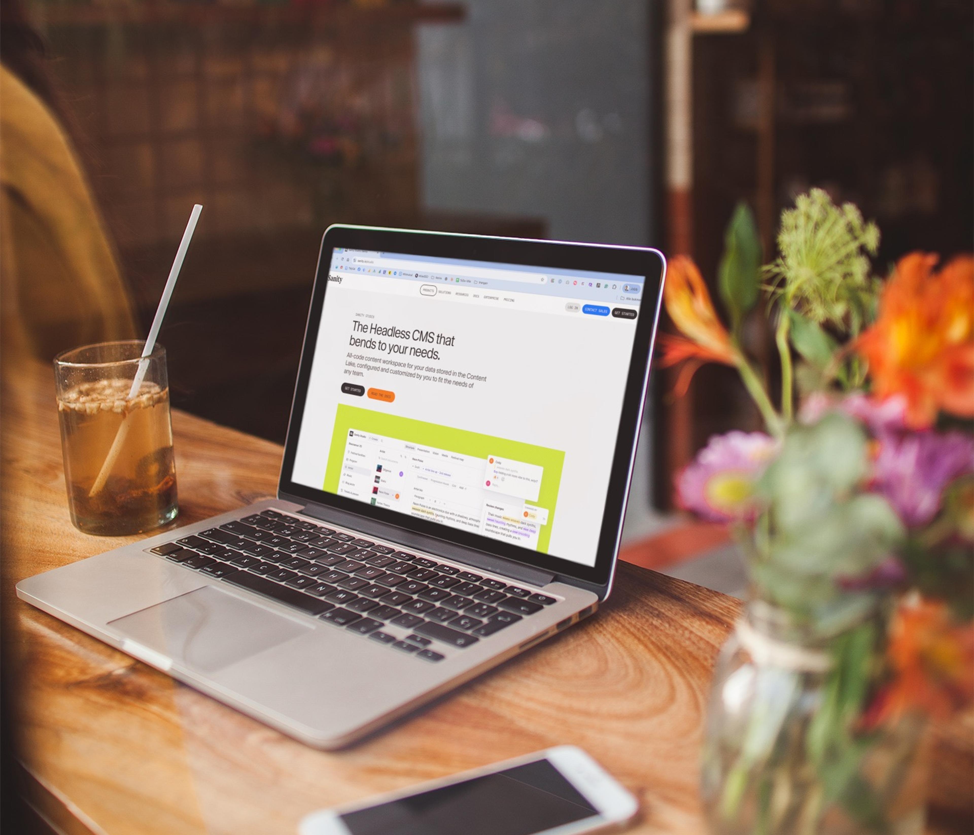 Laptop viser "The Headless CMS that bends to your needs" på tre i kveldslys, med smartphone og blomster på bordet.