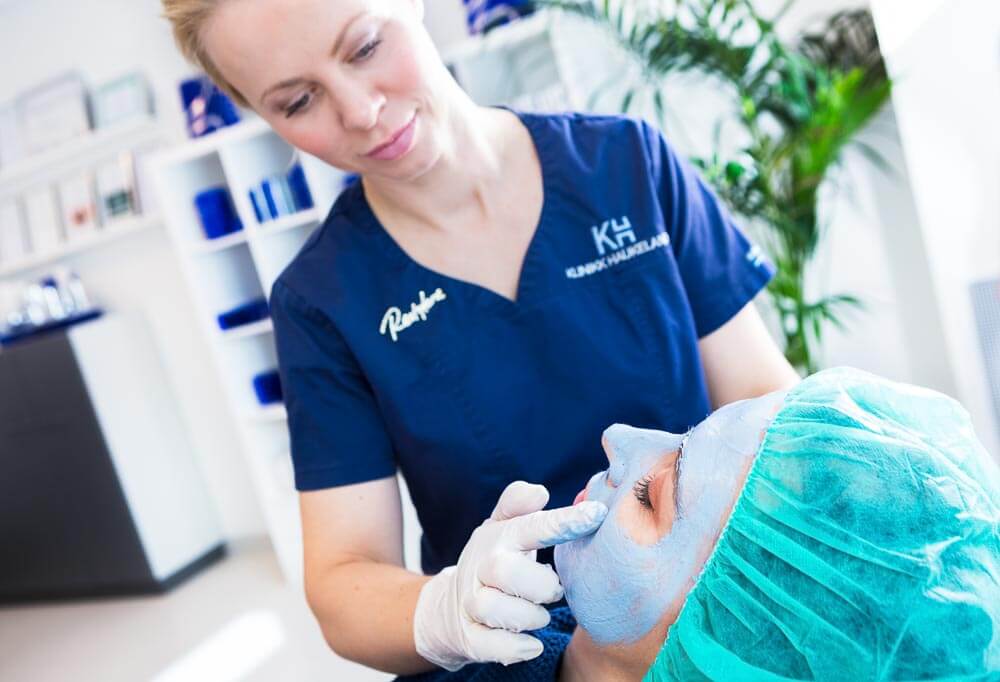 Sykepleier i blå uniform behandler pasient med ansiktsmask på klinikkkontor med planter i bakgrunnen.