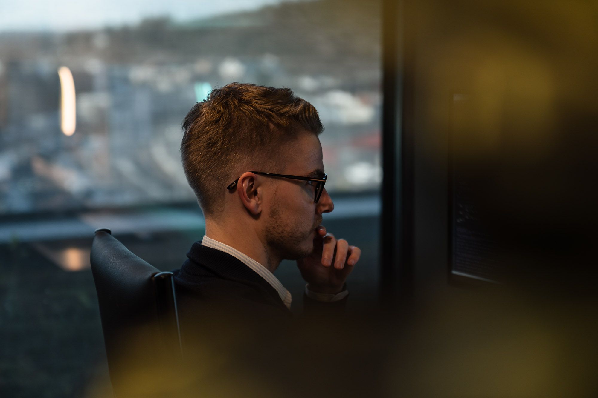 Mann med briller ser ut fra kontorvindu med byutsikt, tenker over arbeidsoppgaver på høyt nivå.