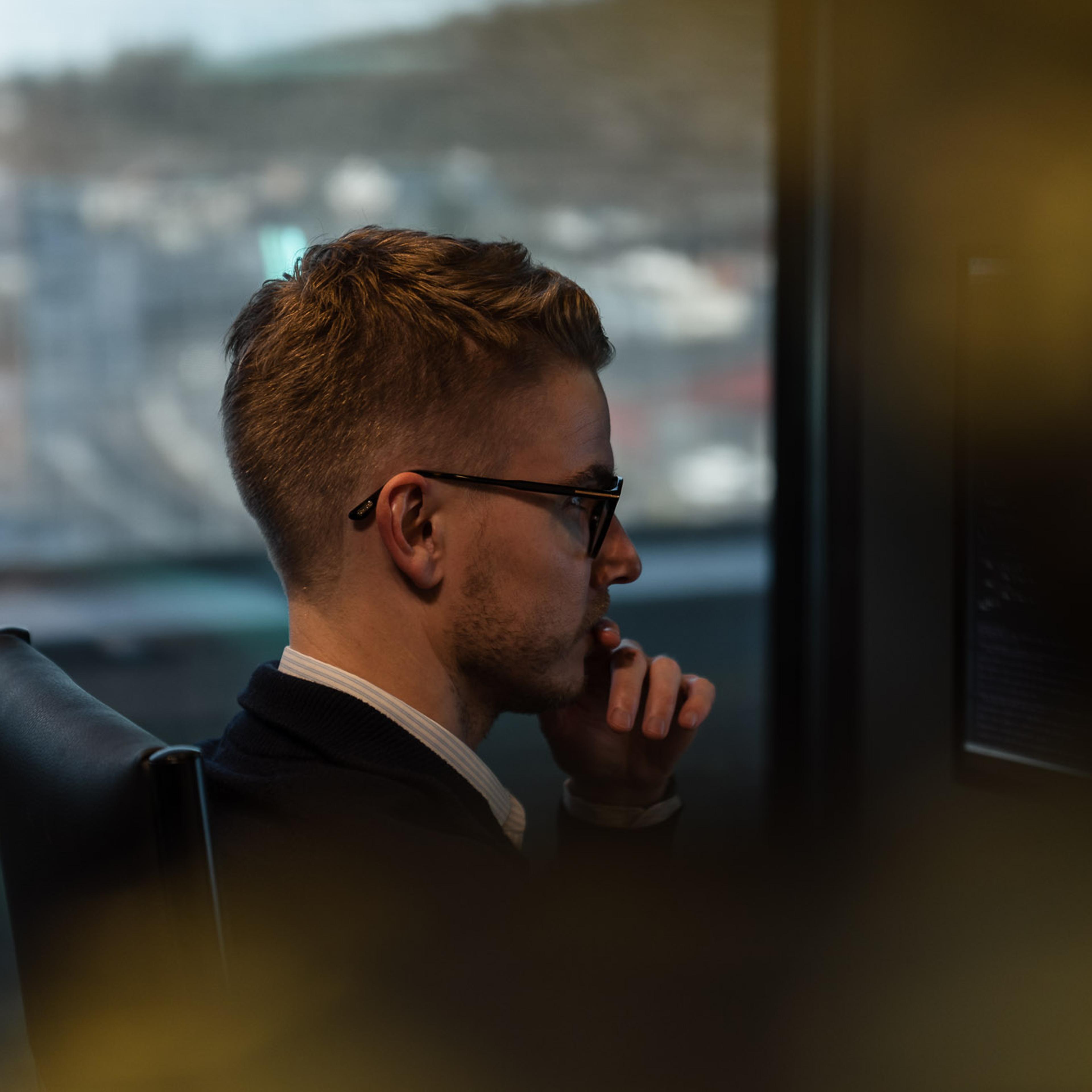 Mann med briller ser ut fra kontorvindu med byutsikt, tenker over arbeidsoppgaver på høyt nivå.