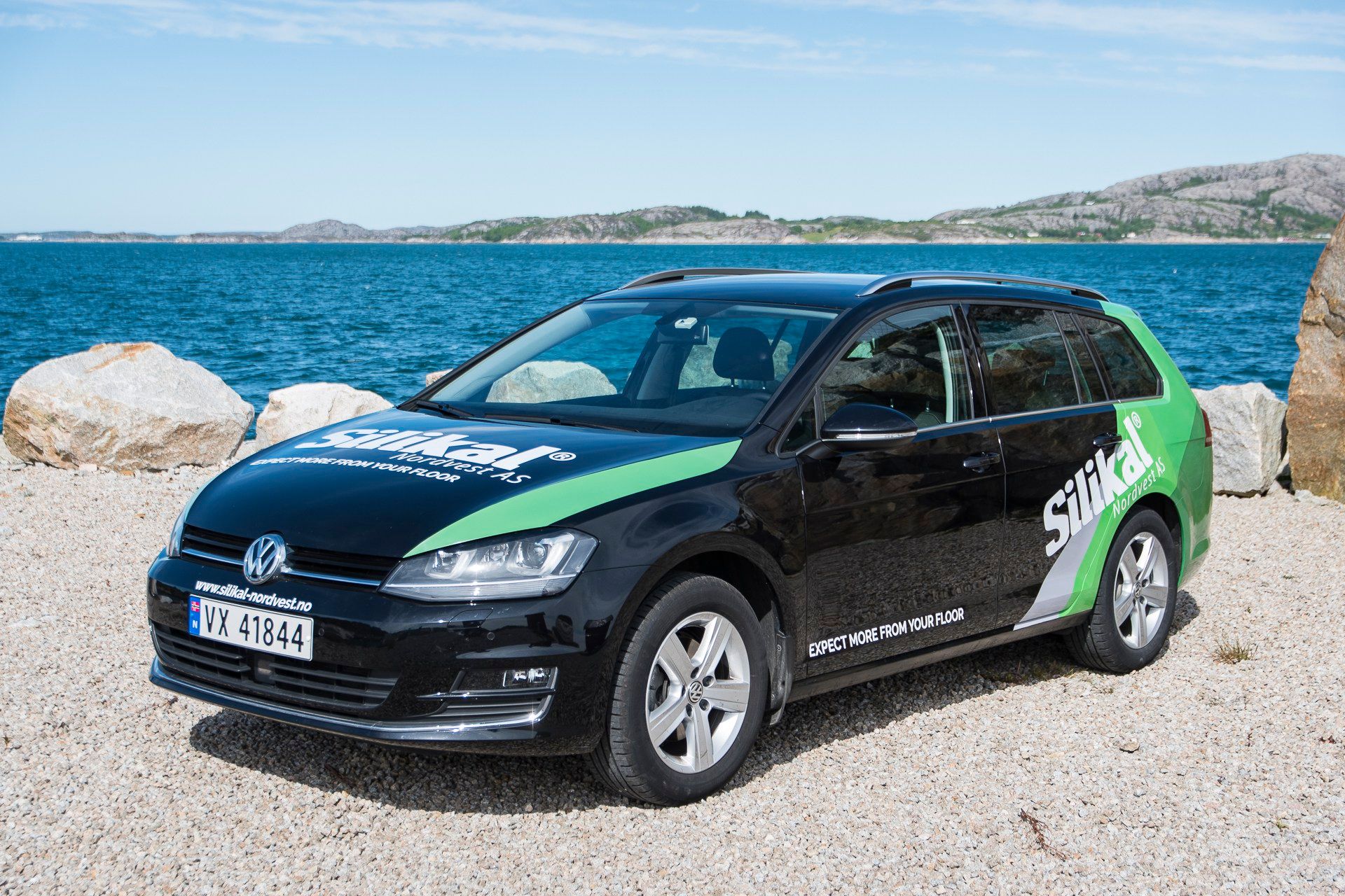 Volkswagen-minibuss med Silikal-folie i svart og grønt, parkert ved strand med blå hav i bakgrunnen.