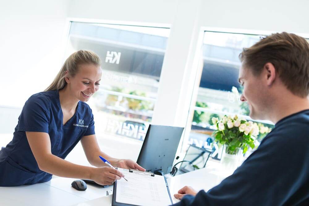 Helsepersonell i blå uniform skriver notater mens hun møter pasient ved skranke på klinikk.