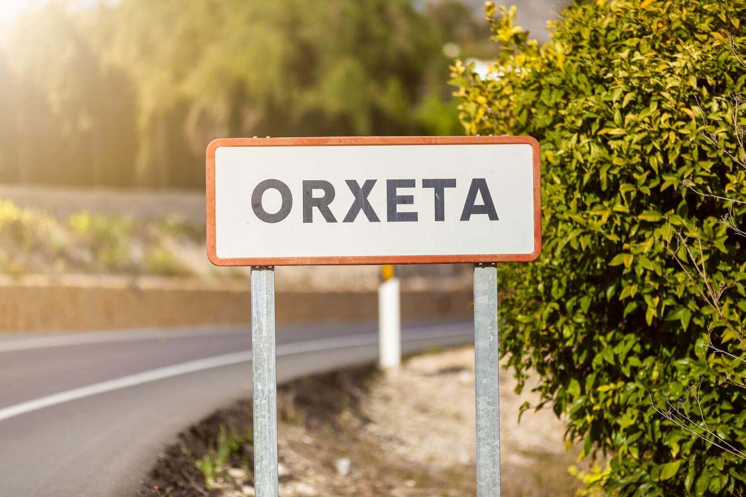 Veiskilt med teksten ORXETA ved en landsvei omgitt av grønn vegetasjon og solskinn.