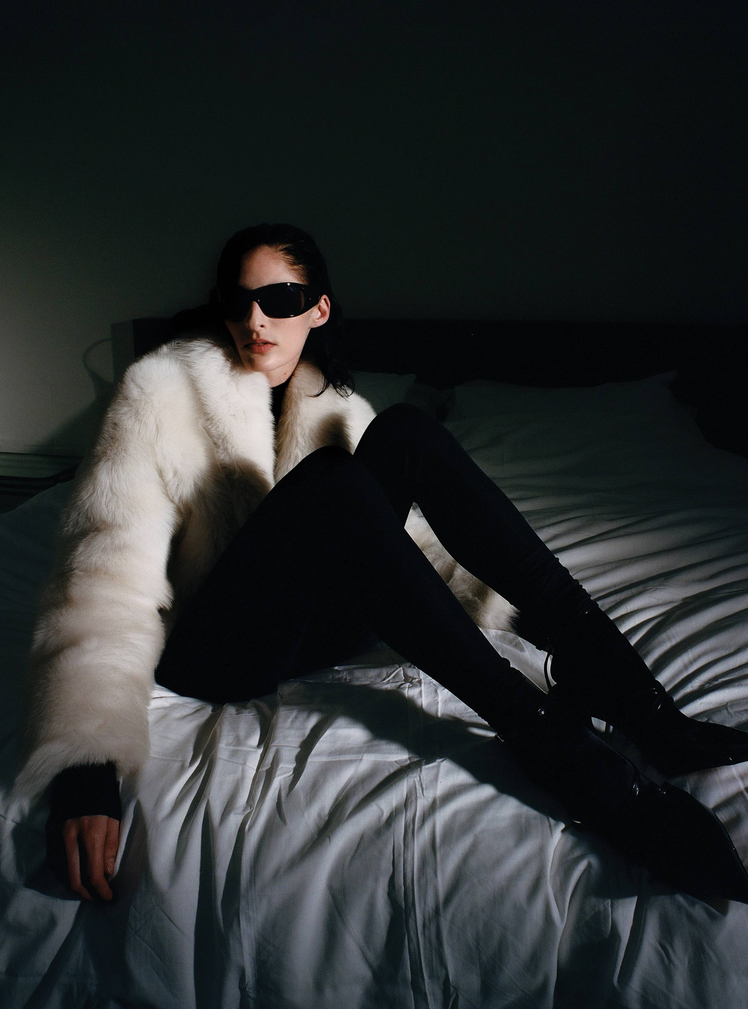 A model sitting on a bed wearing the Soiree Cloud fluffy shearling coat