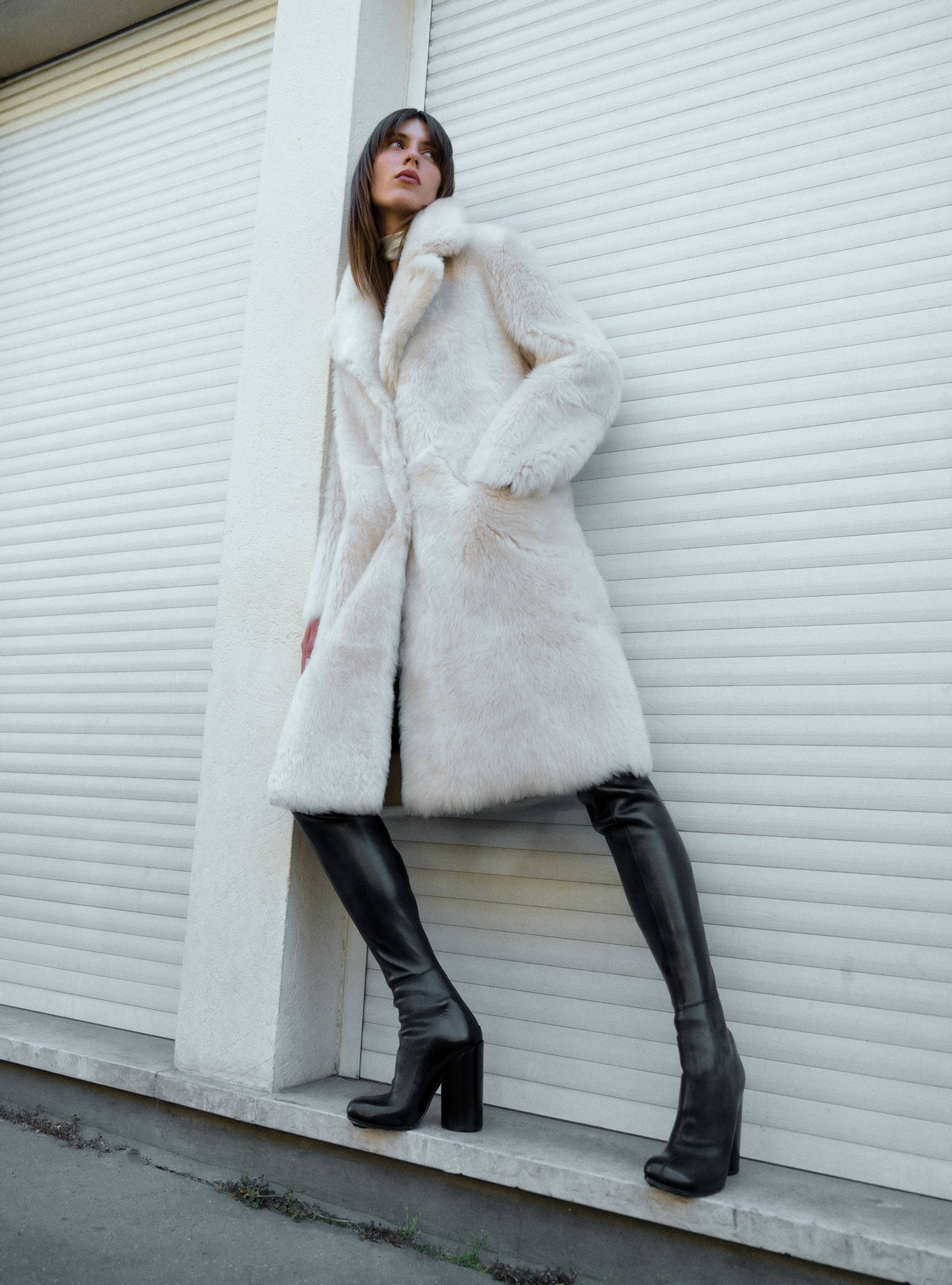 A model leaning against a white wall in the Evita Ivory Apres Ski Shearling Choat