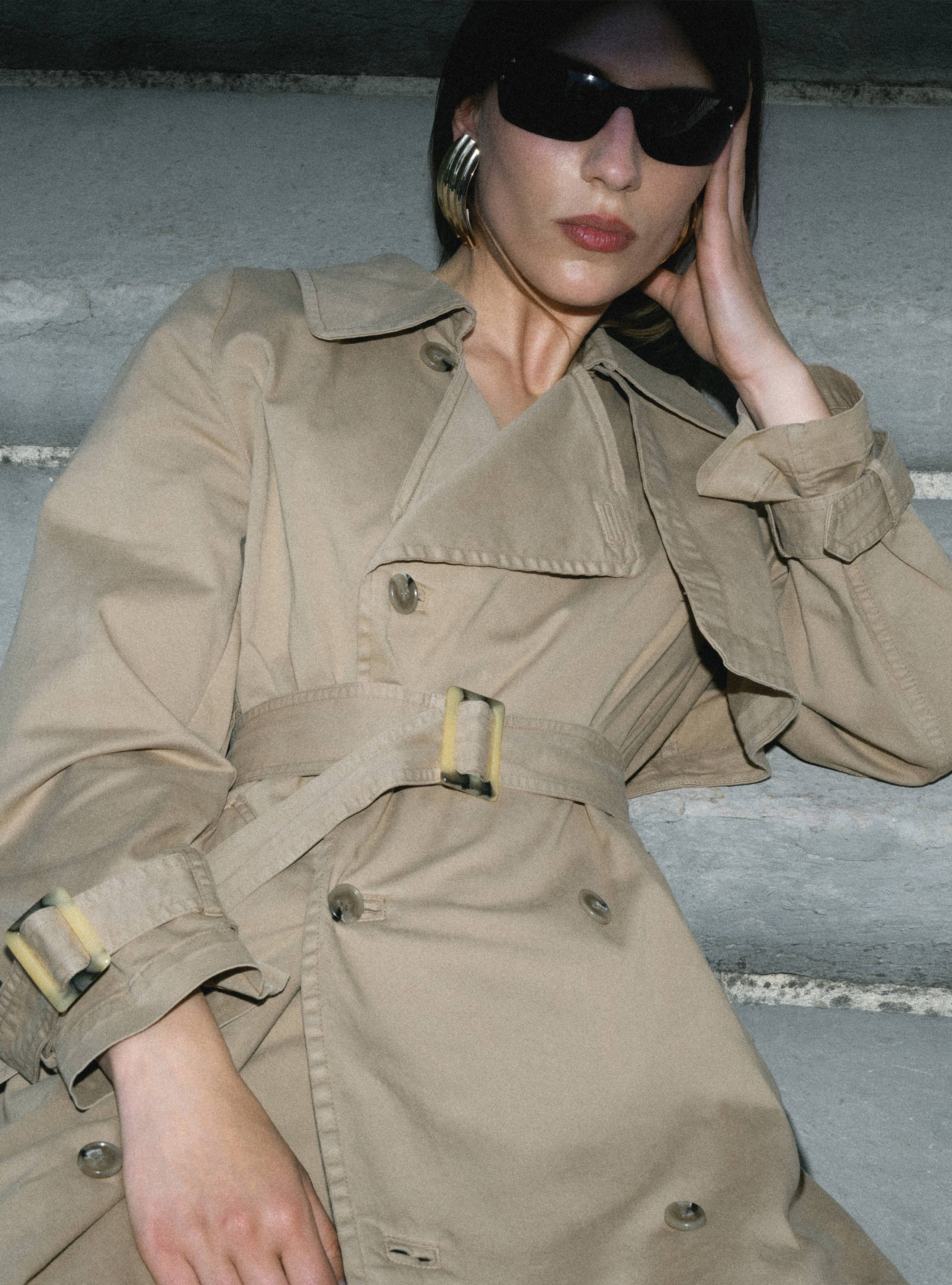 A model sitting on a staircase wearing the Harriet Canvas Khaki trench coat