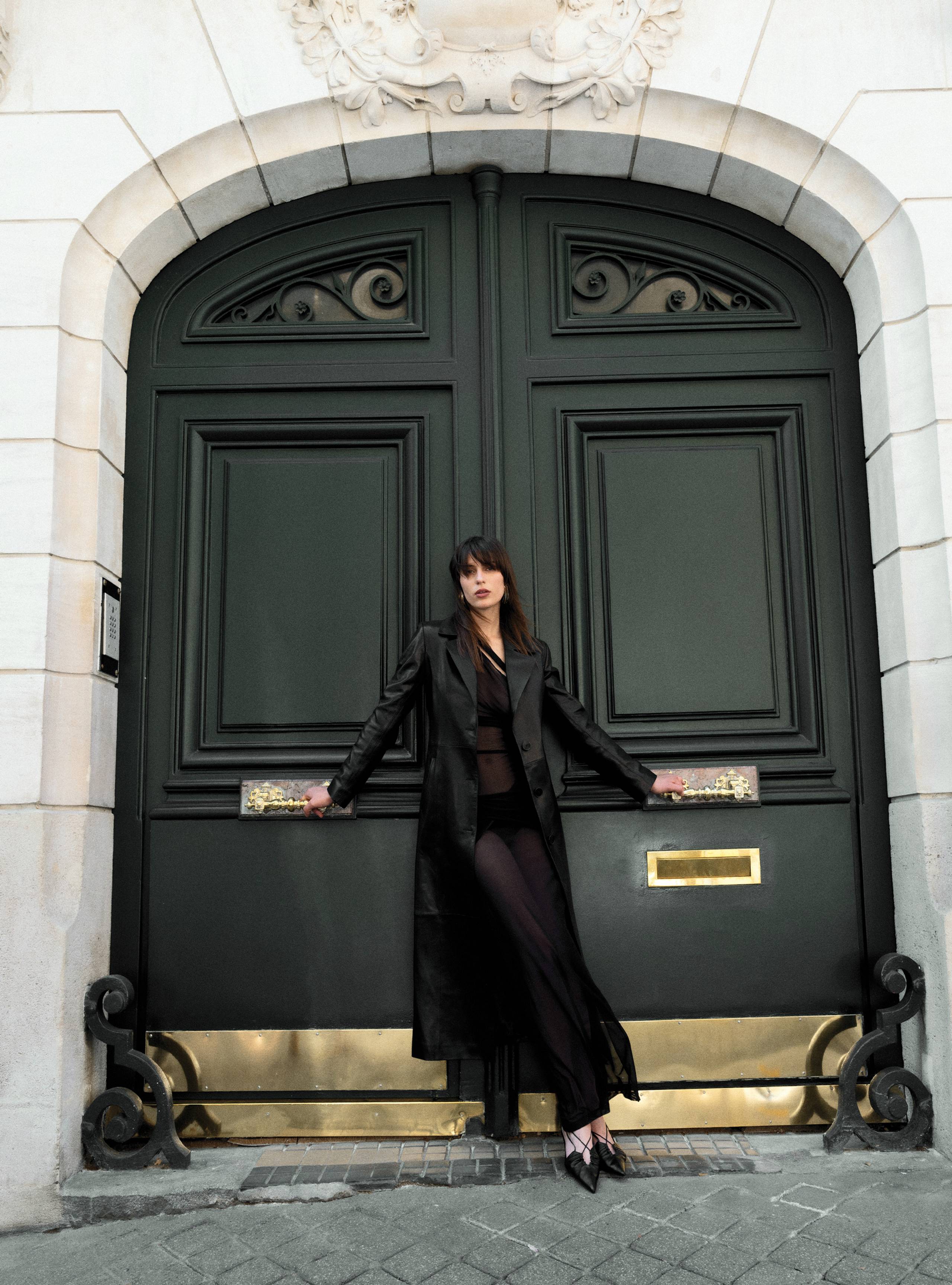A model standing in front of a door wearing the Tamara Black long coat