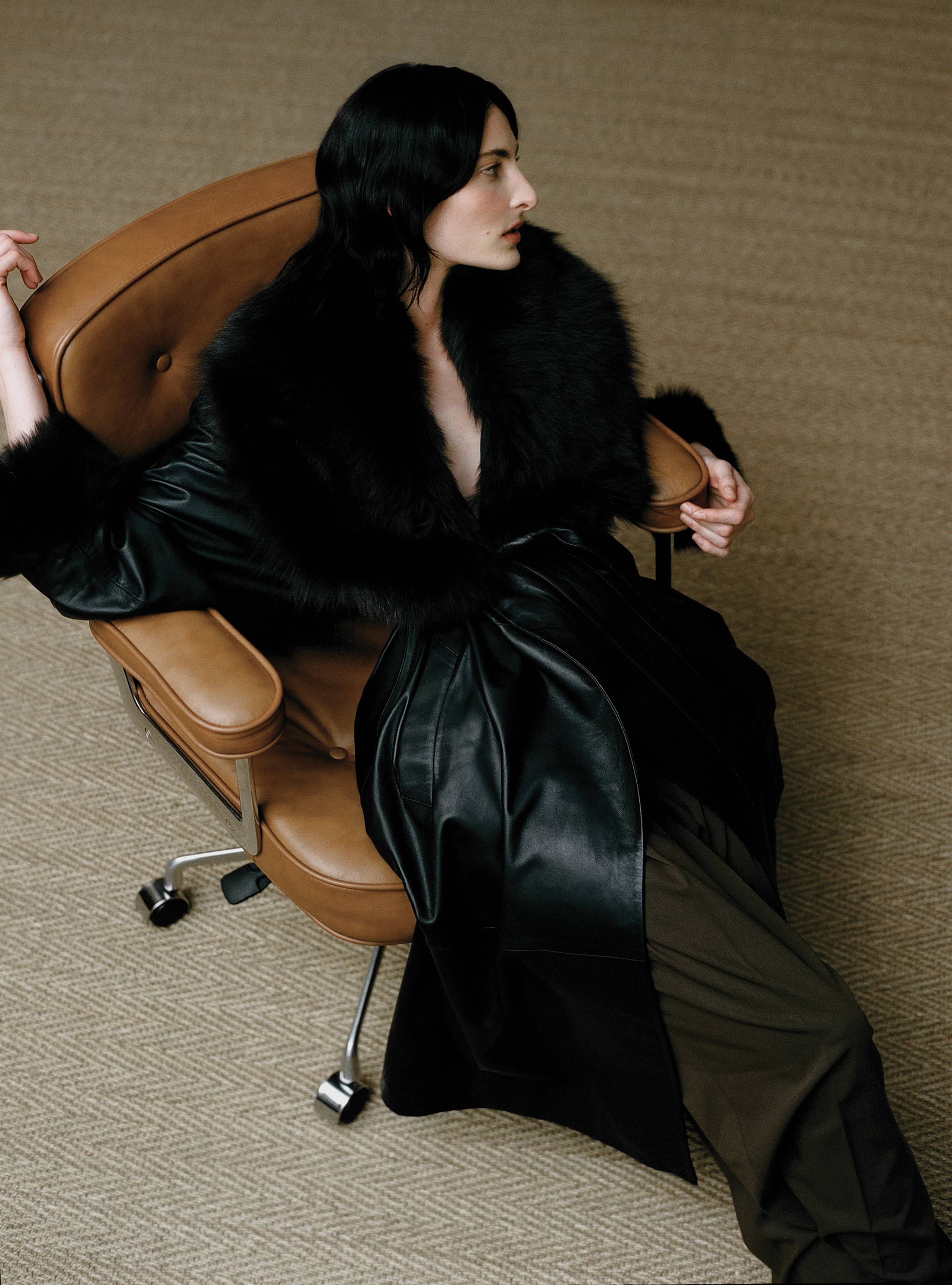 A model sitting on a chair wearing the Freja Black Dark Chocolate mid-length leather trench