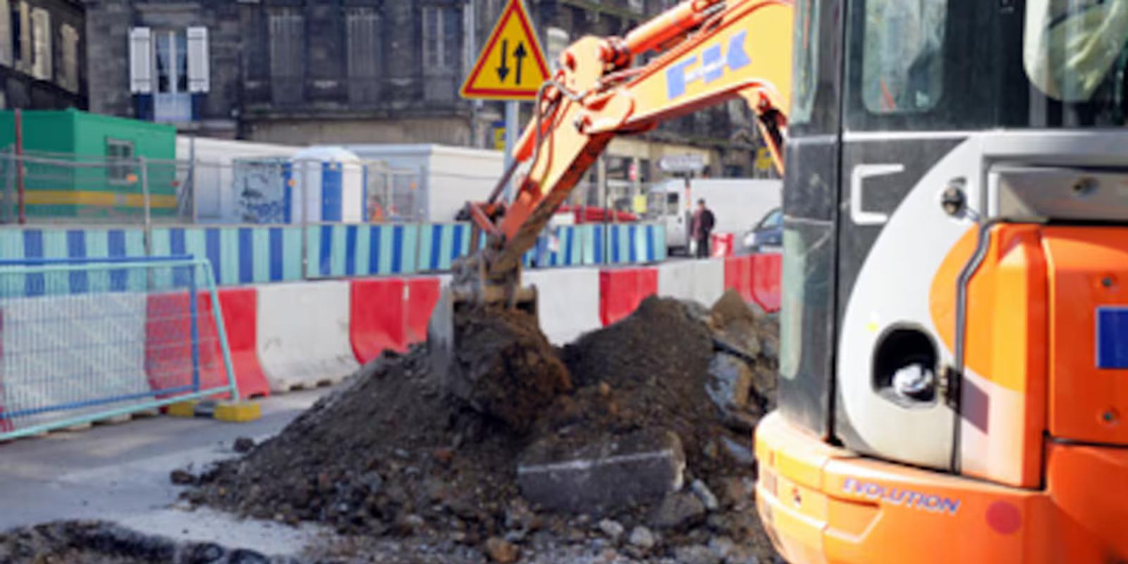 Chantier de travaux publics à l'arrêt devant un tableau de variation des prix des matières premières, illustrant l'imprévision en marché public.