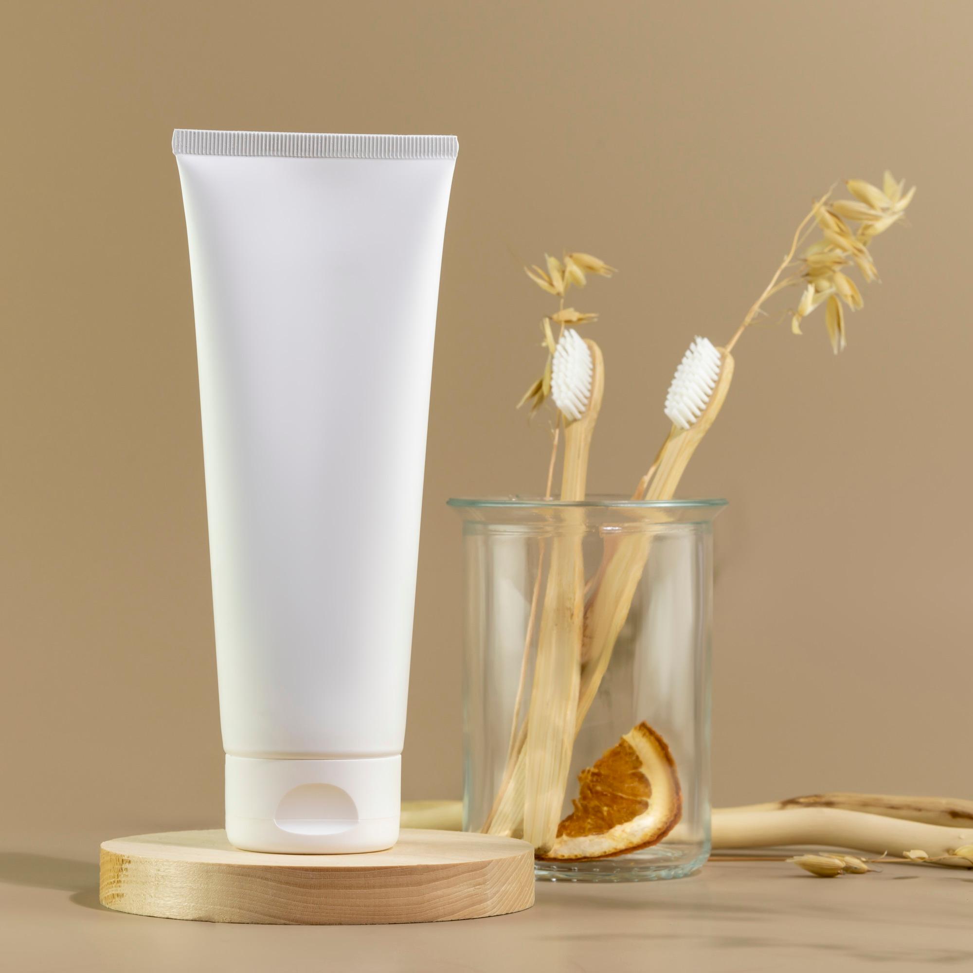 Tube med tannkrem og tannbørster i et glass mot sandfarget bakgrunn