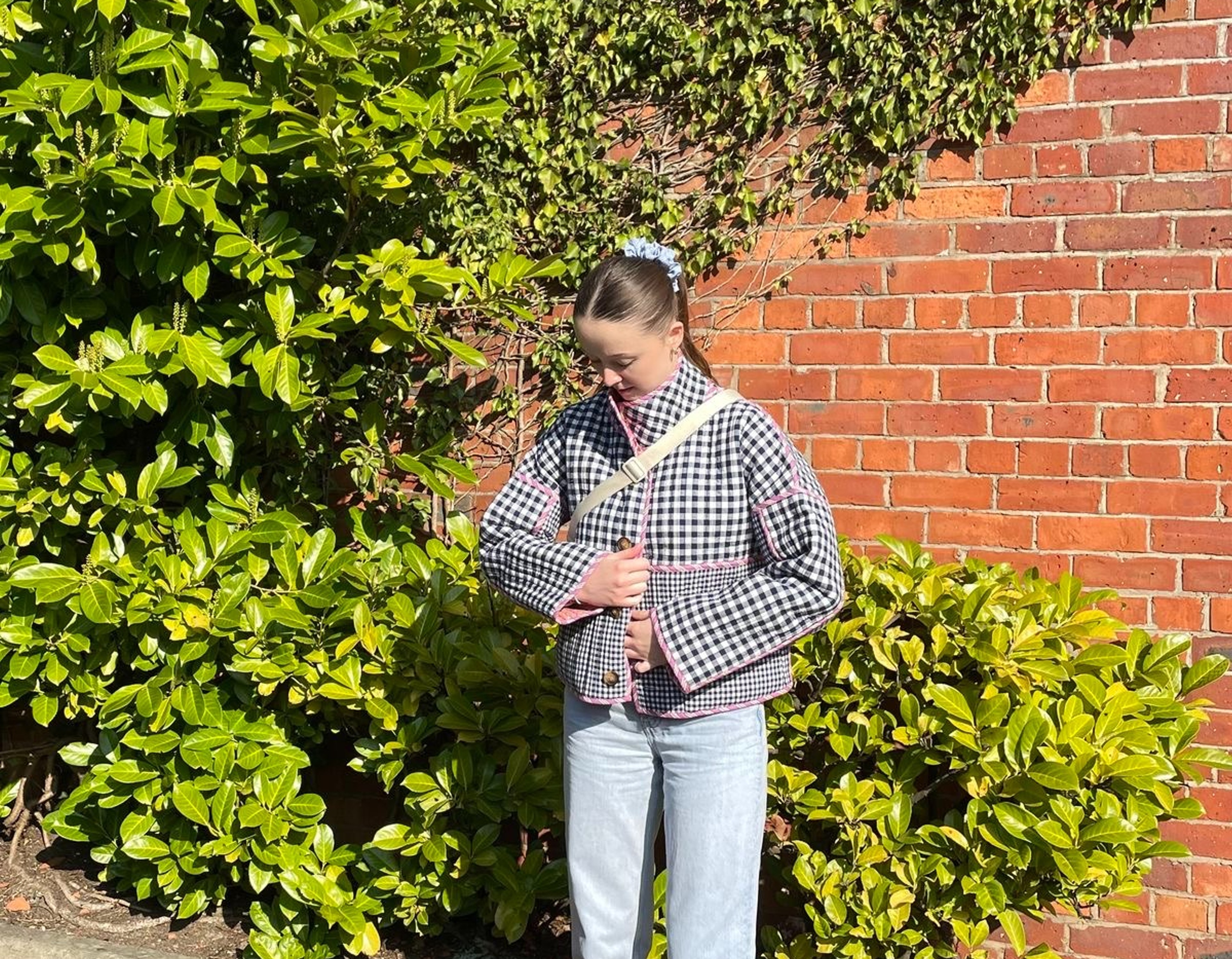 A girl stood in front of a brick wall with green plants wearing jeans and navy checked jacket