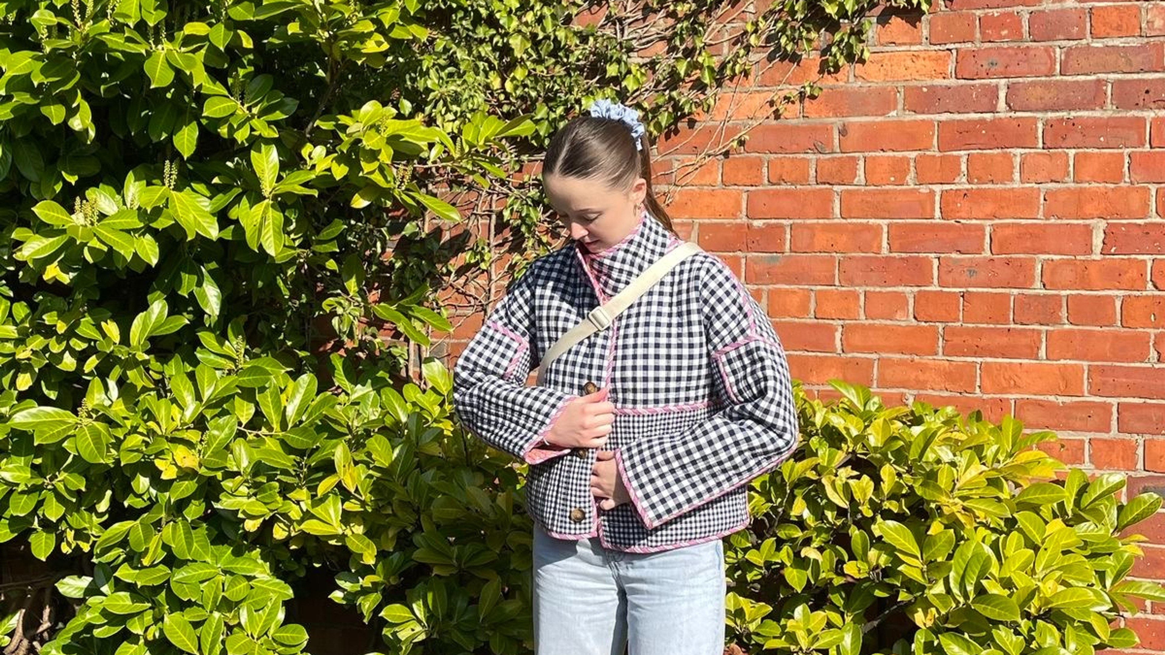 A girl stood in front of a brick wall with green plants wearing jeans and navy checked jacket