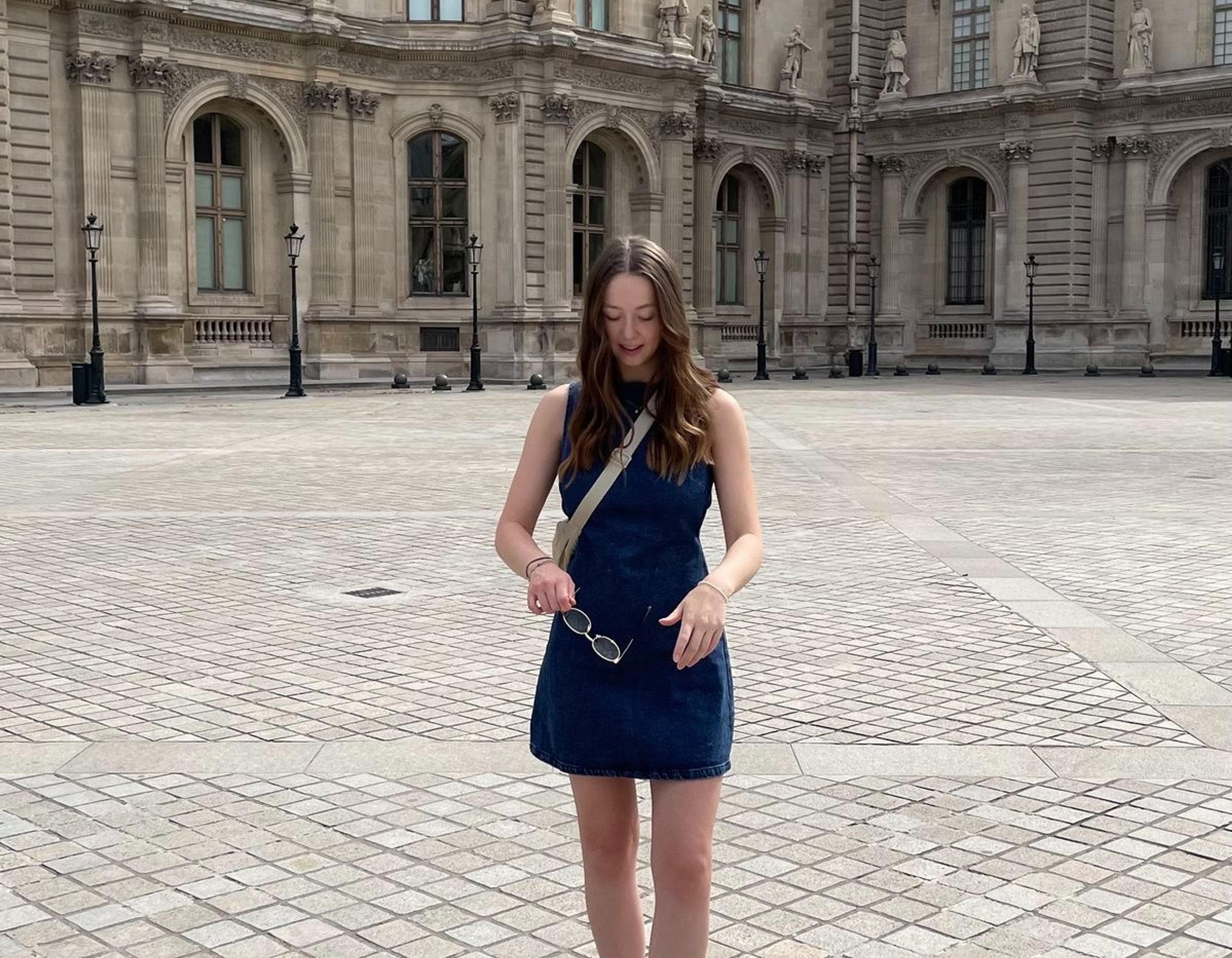 A girl in a denim dress and red shoes in front of the Louvre.