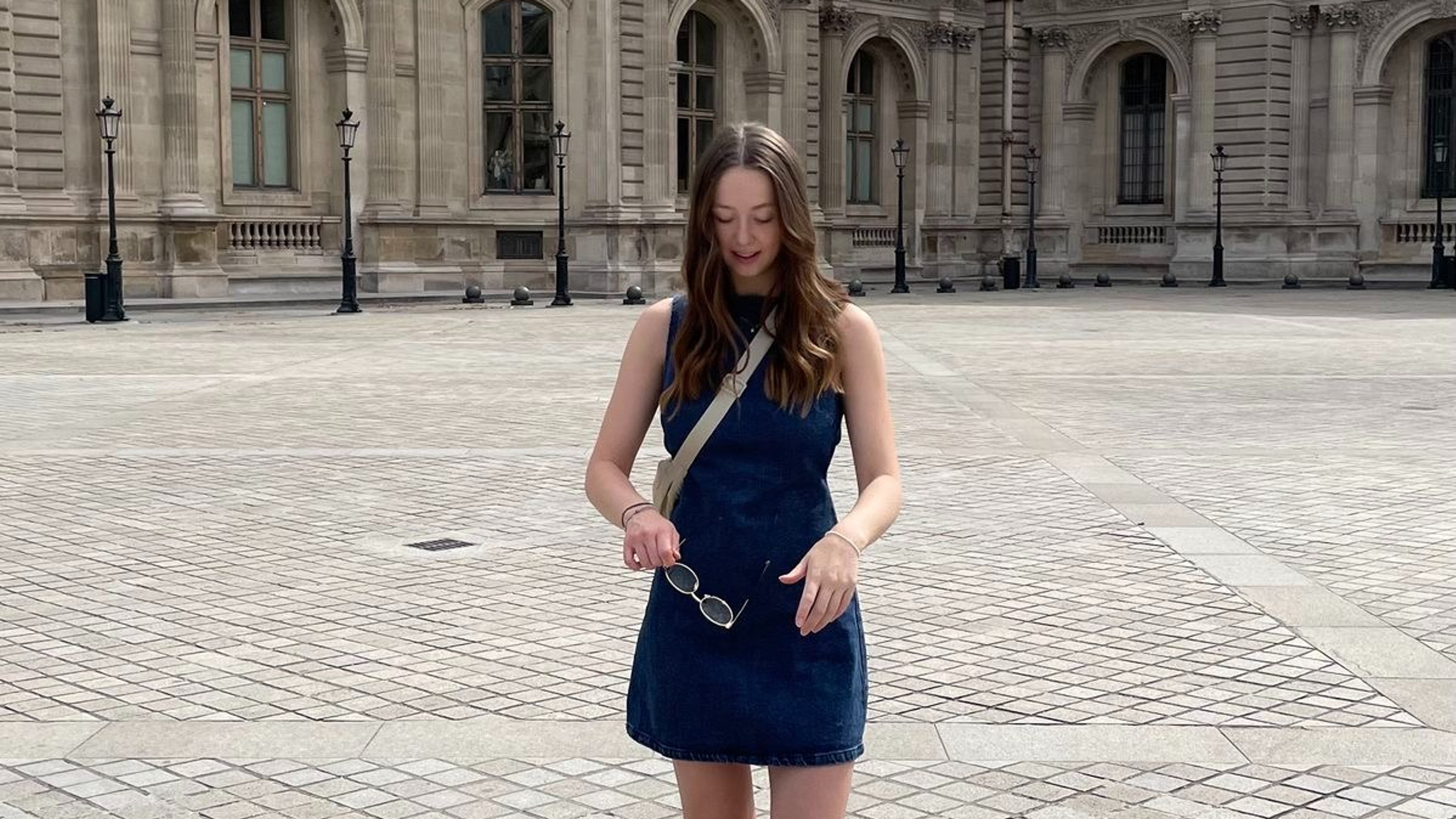 A girl in a denim dress and red shoes in front of the Louvre.