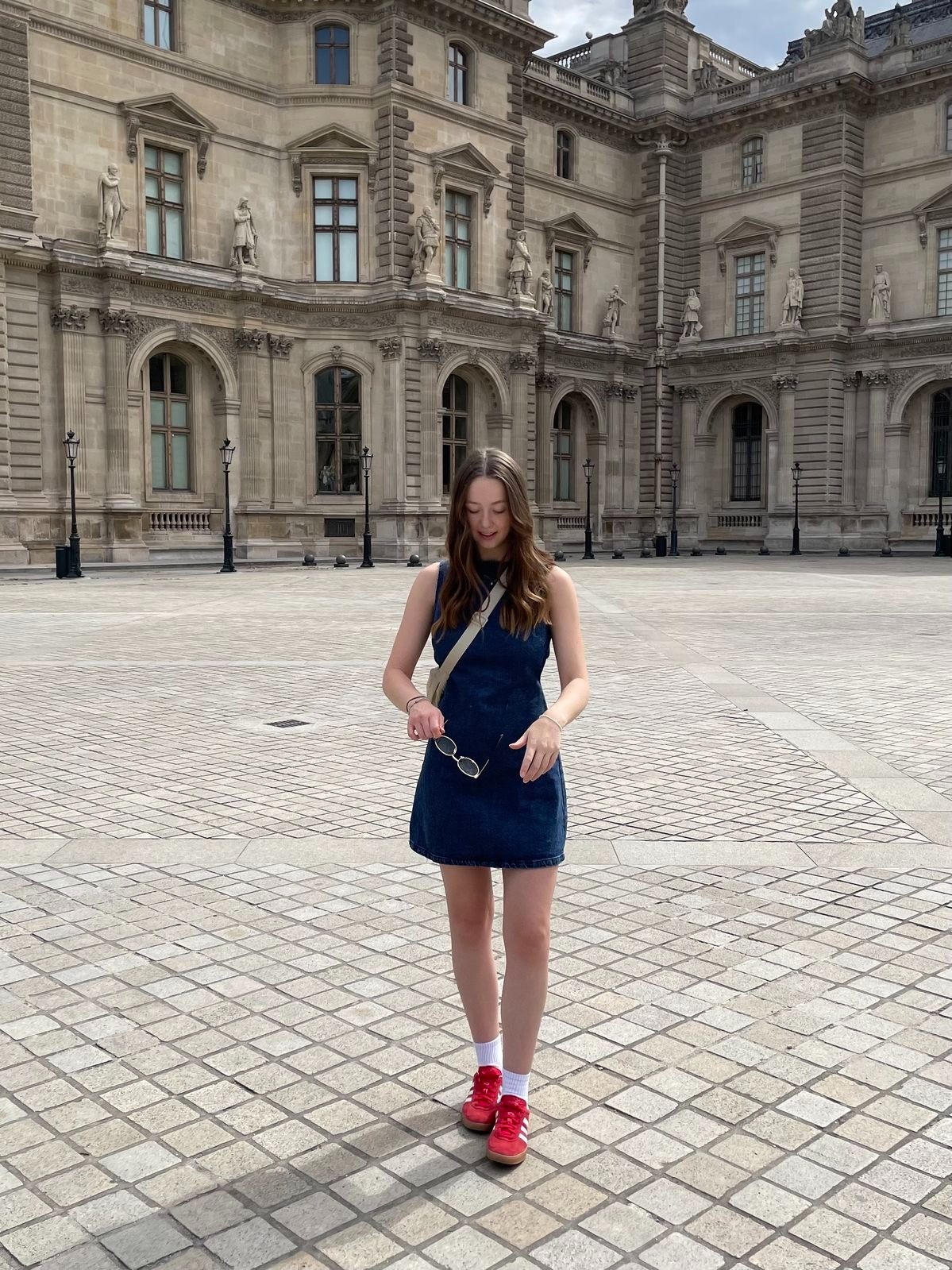 A girl in a denim dress and red shoes in front of the Louvre.