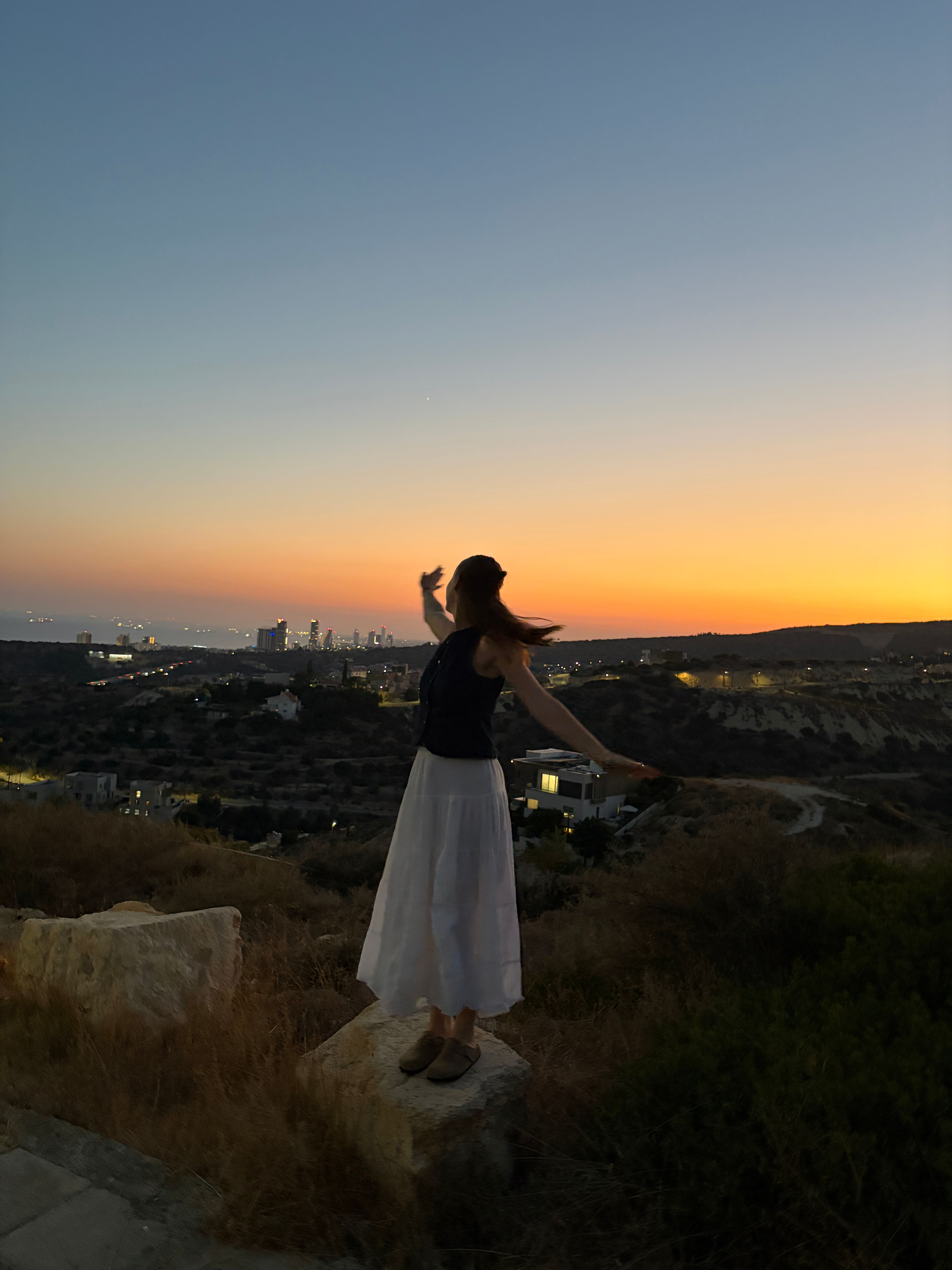 Girl stood on a mountain at sunset looking out at the city horizon 