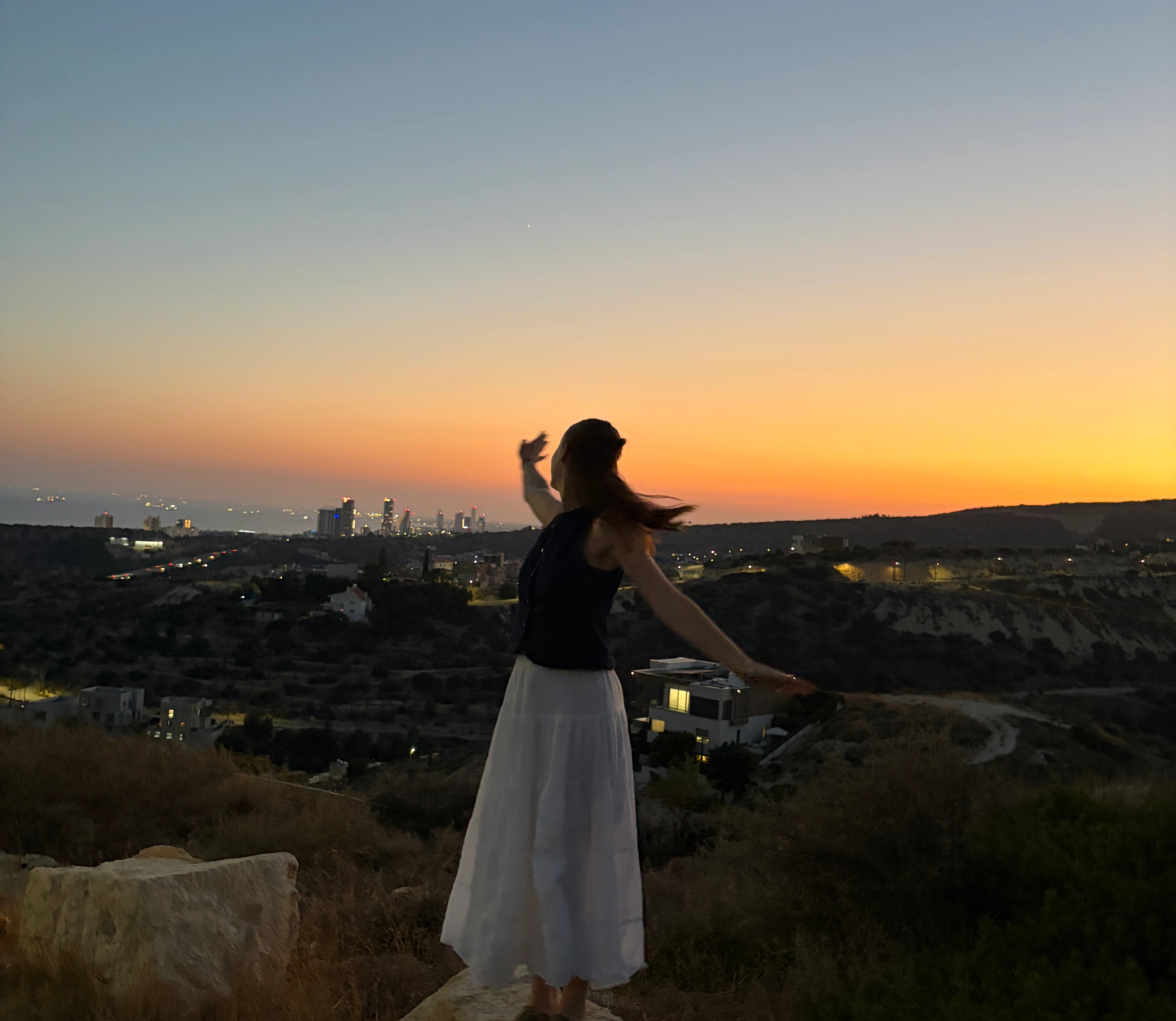 Girl stood on a mountain at sunset looking out at the city horizon