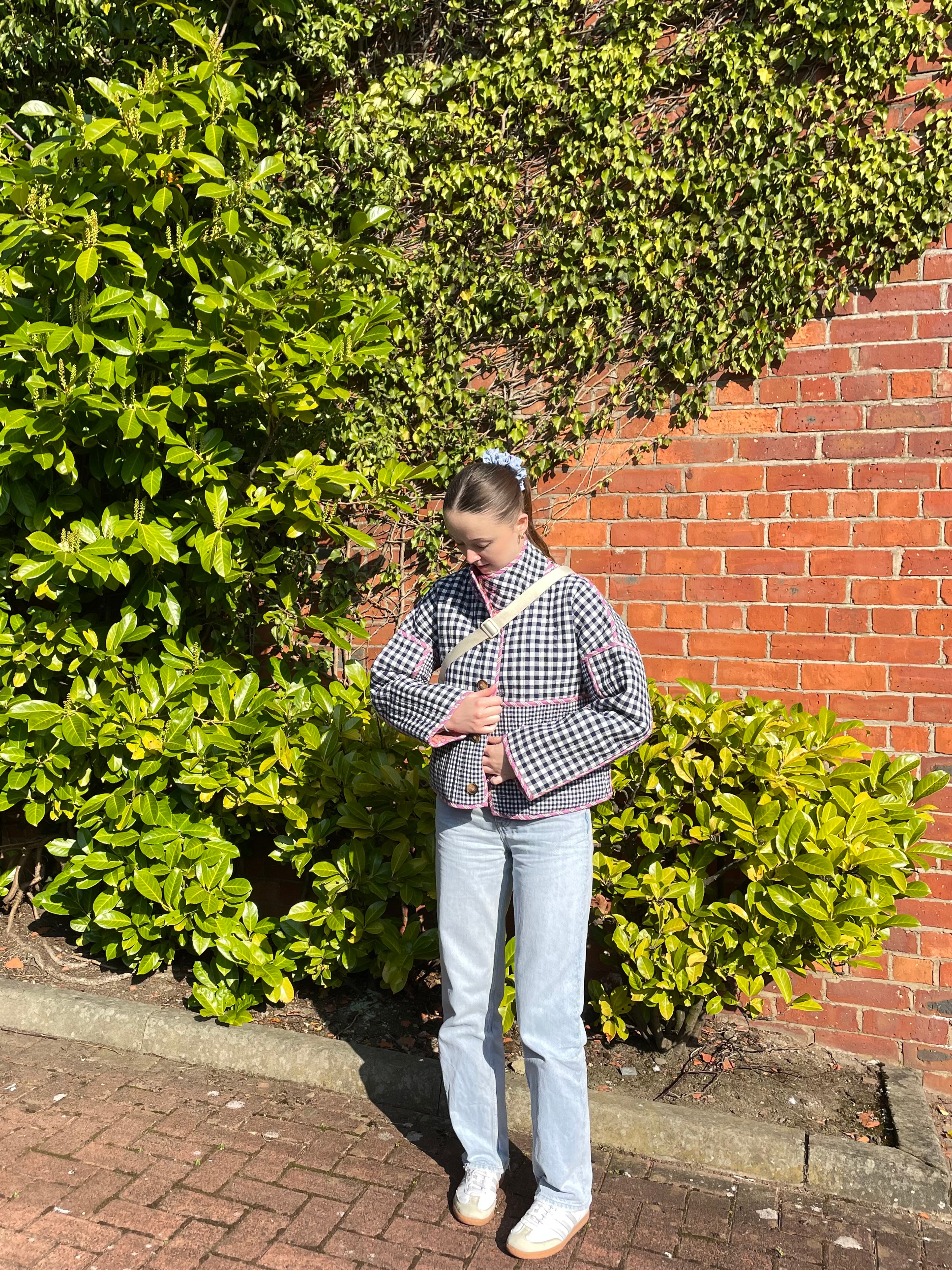 A girl stood in front of a brick wall with green plants wearing jeans and navy checked jacket