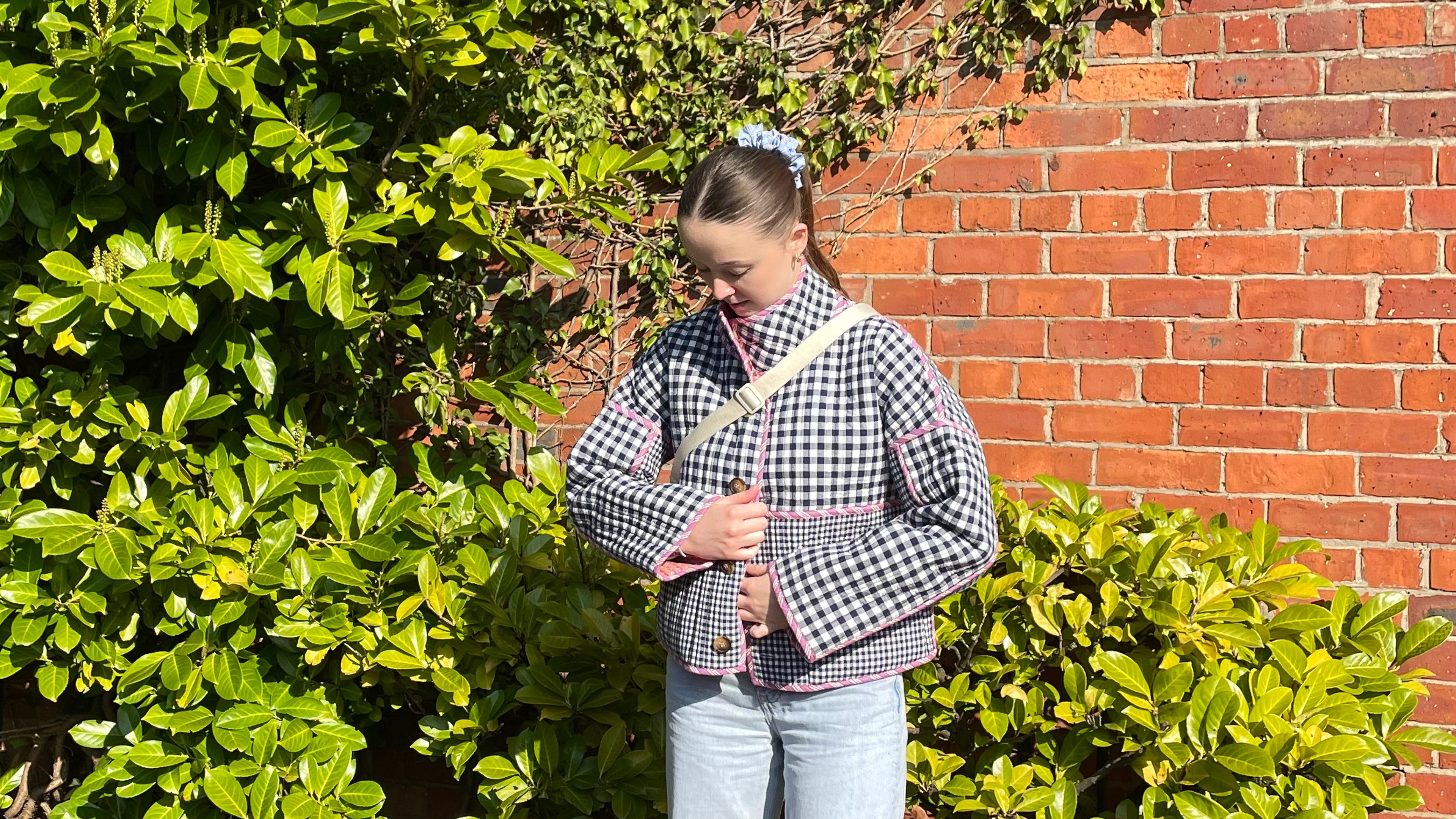 A girl stood in front of a brick wall with green plants wearing jeans and navy checked jacket