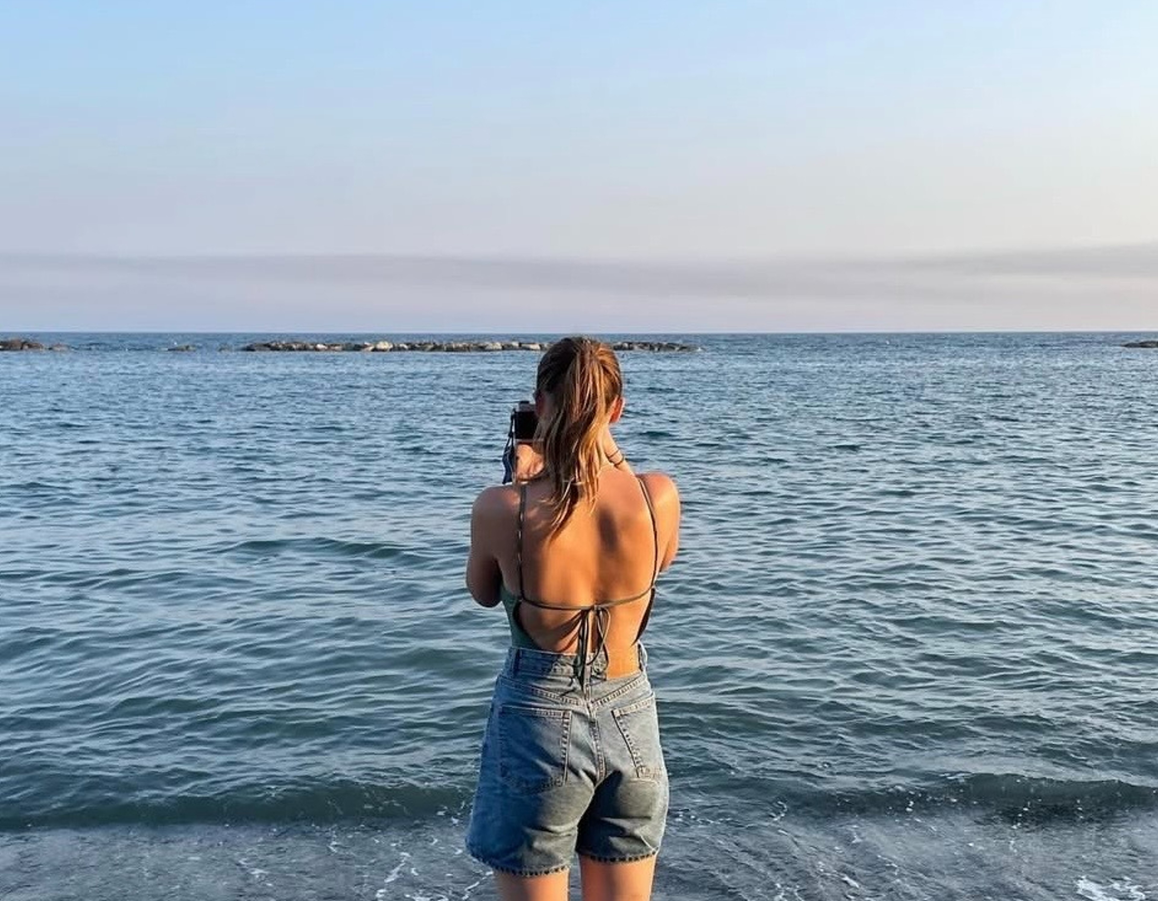 A girl stood on the beach looking out to sea holding a film camera and taking a picture.